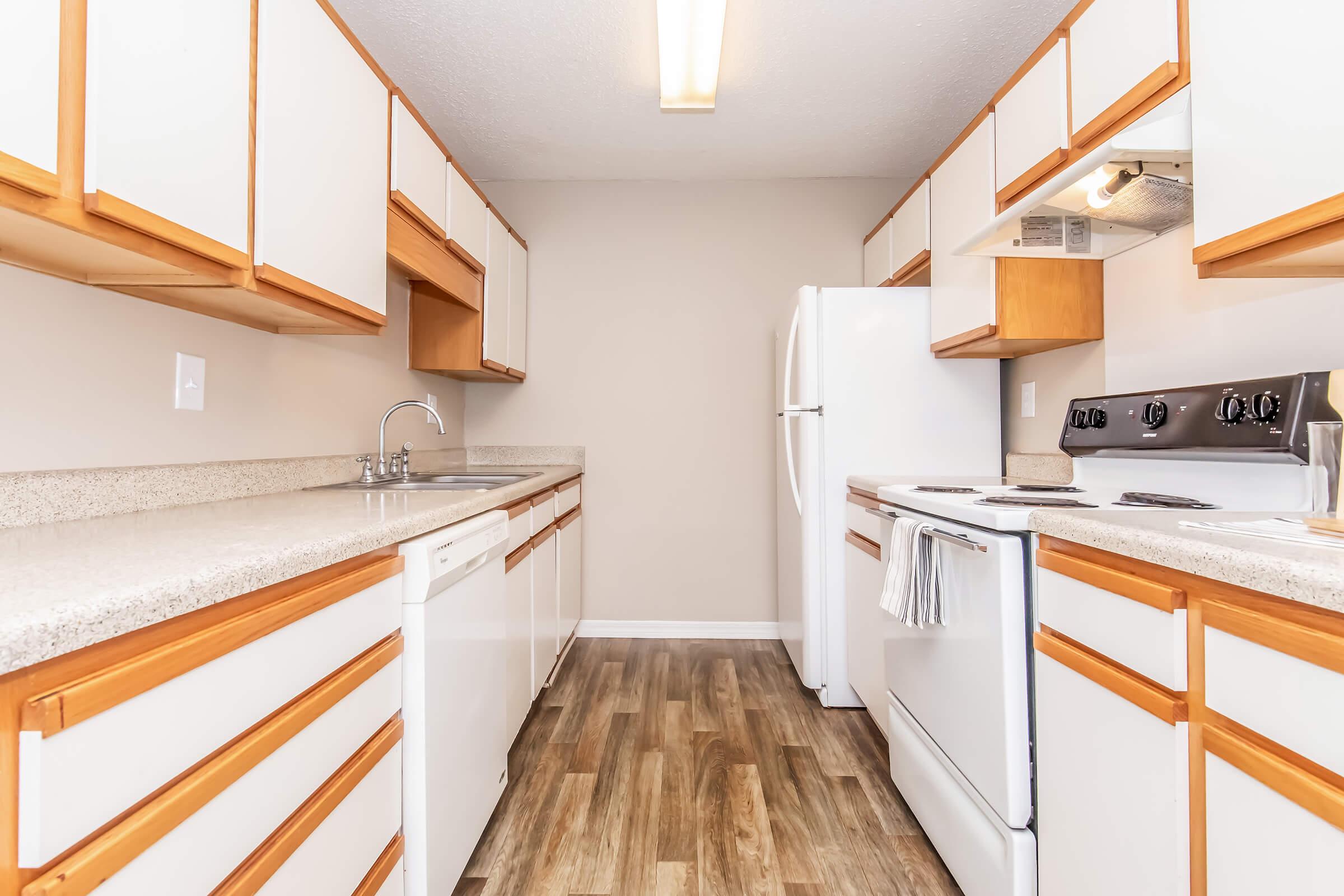 A modern kitchen featuring light wood cabinets, a white countertop, a stainless-steel sink, and a dishwasher. There is a white refrigerator and an oven with a stovetop. The flooring is a wood-like laminate, and the walls are painted a neutral color, creating a bright and spacious atmosphere.