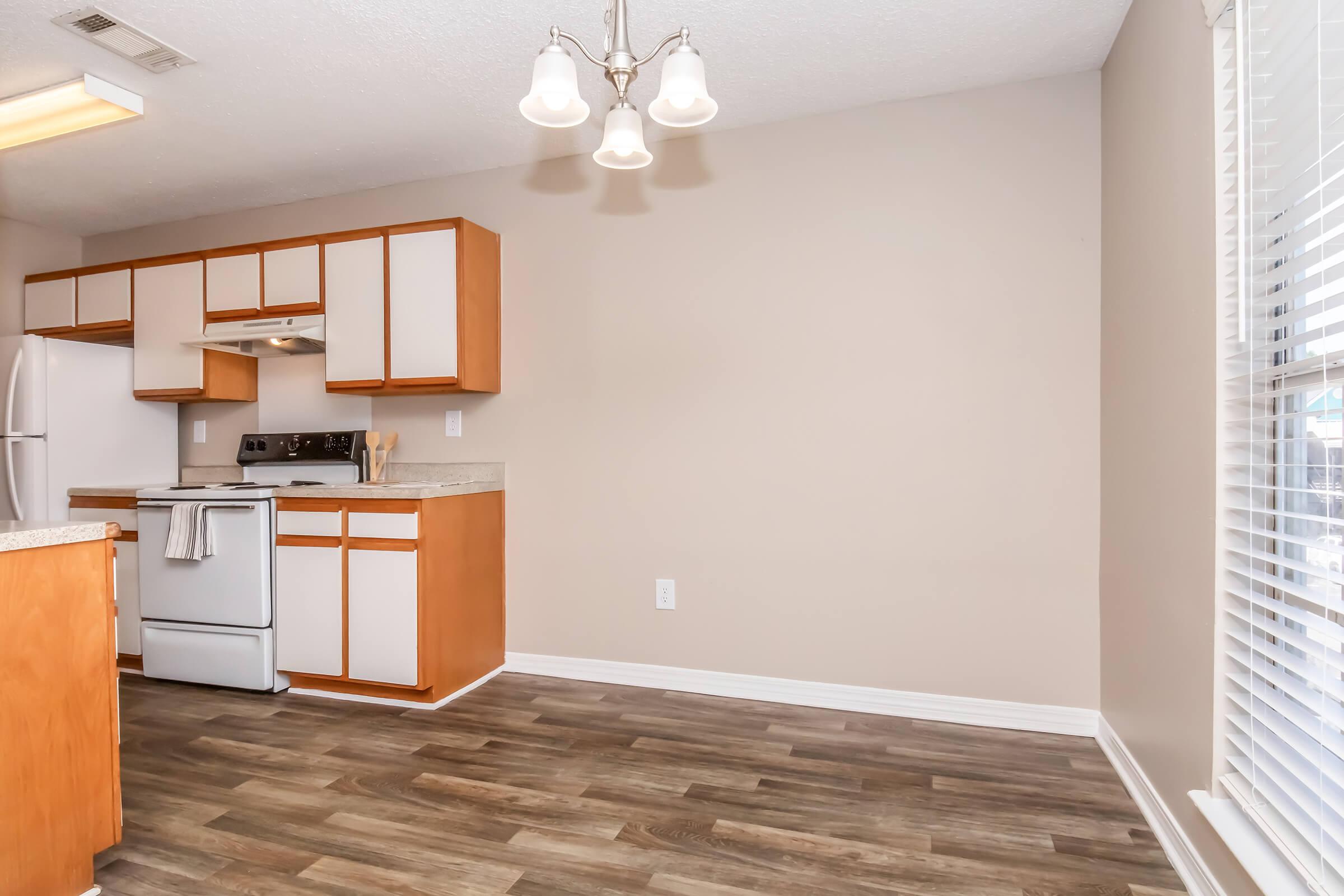 A modern kitchen featuring wooden cabinets, a white refrigerator, and an electric stove. The walls are a soft beige, and the floor has a warm wood-like finish. Bright natural light enters through a window with blinds, and three pendant lights hang from the ceiling, illuminating the space.