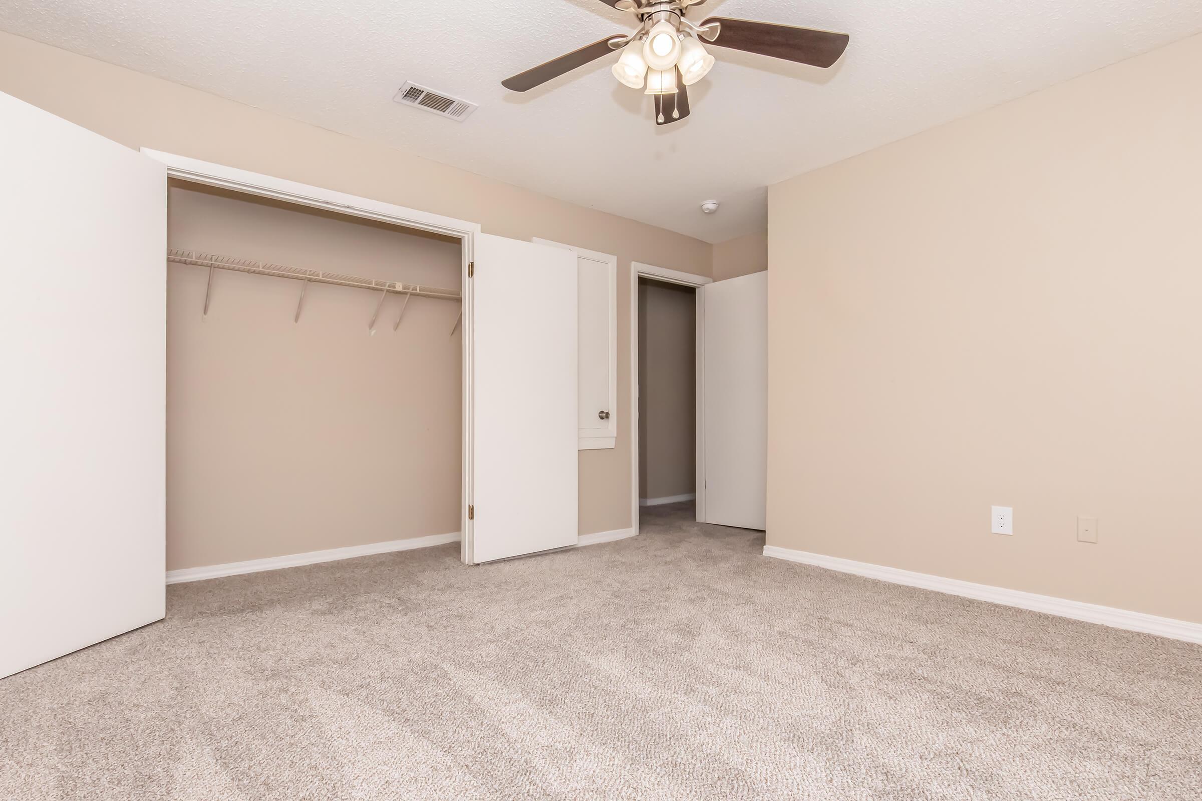 A cozy, empty bedroom featuring light beige walls, a ceiling fan, and plush carpet. There are two open closet doors on the left, leading to a spacious closet, and a doorway visible on the right. The room is well-lit and ready for personalization.