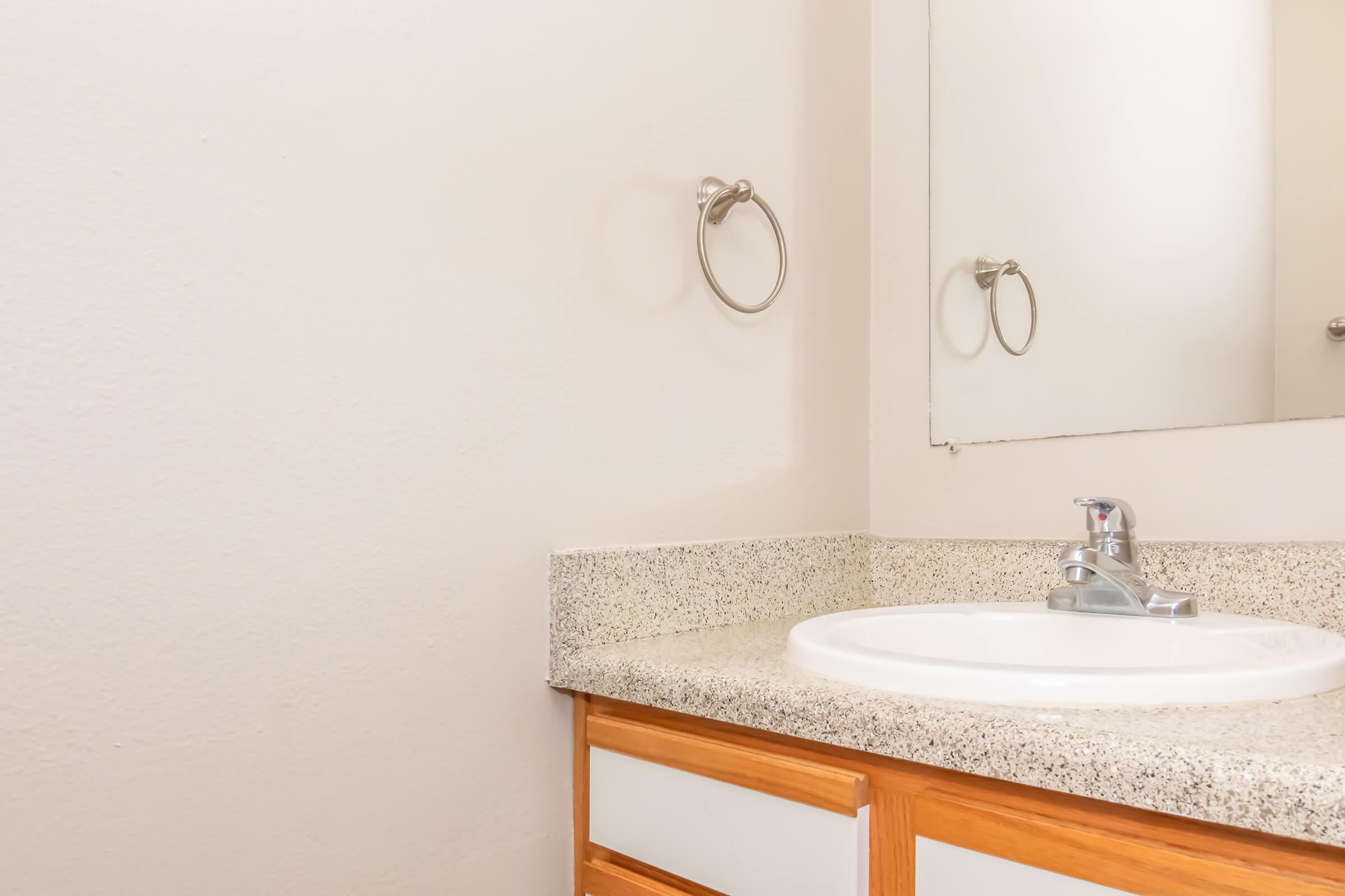 A simple bathroom sink area featuring a white basin on a light-colored speckled countertop, with wooden cabinet drawers beneath. A wall-mounted mirror reflects part of the space, and a chrome towel ring is mounted on the wall. The background is a plain beige wall, creating a clean and minimalistic look.