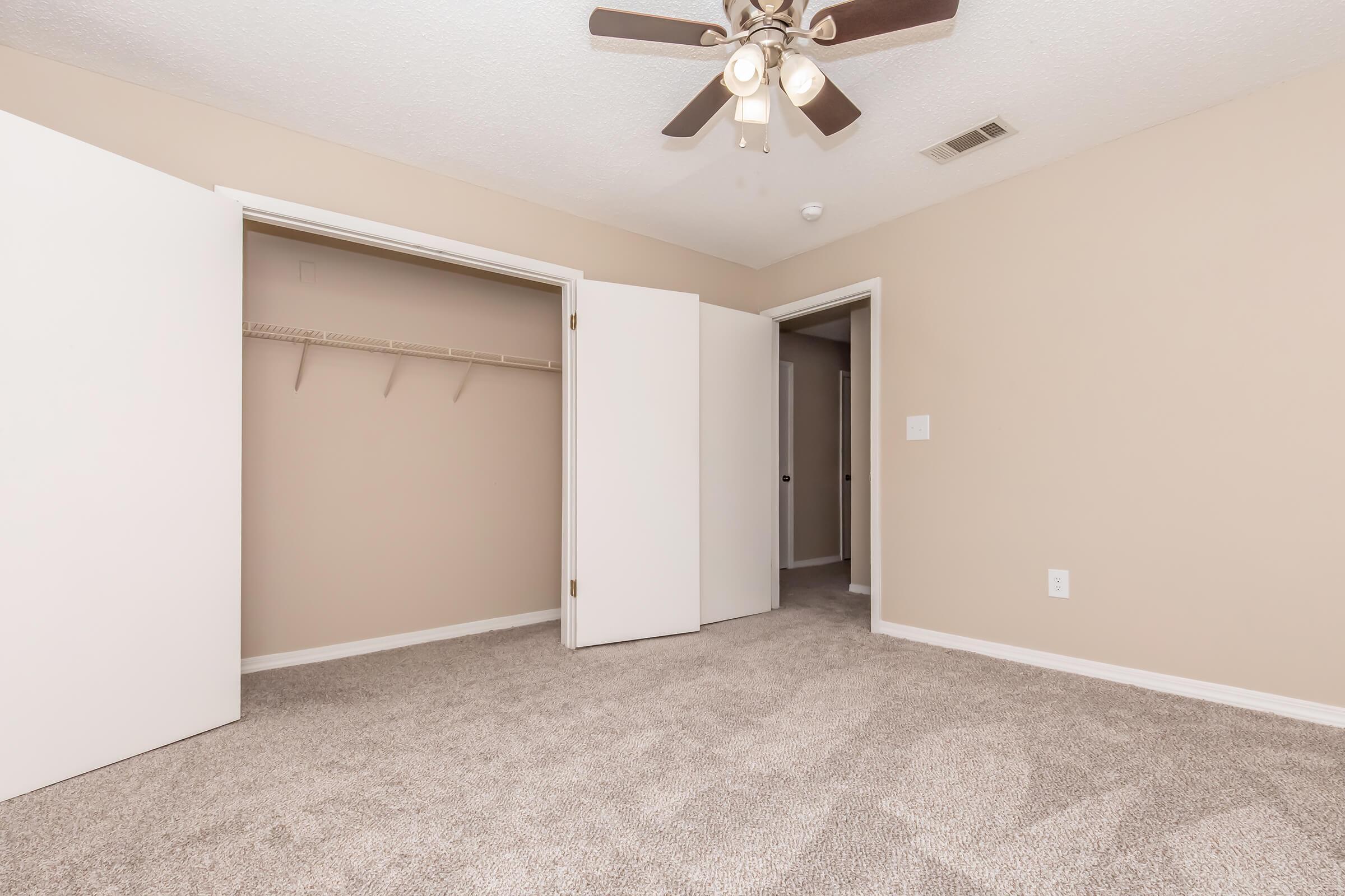 A spacious, well-lit bedroom featuring light beige walls, soft carpet, and a ceiling fan. There are two white doors leading to a closet and another room. The room has a neutral aesthetic, creating a calm atmosphere.