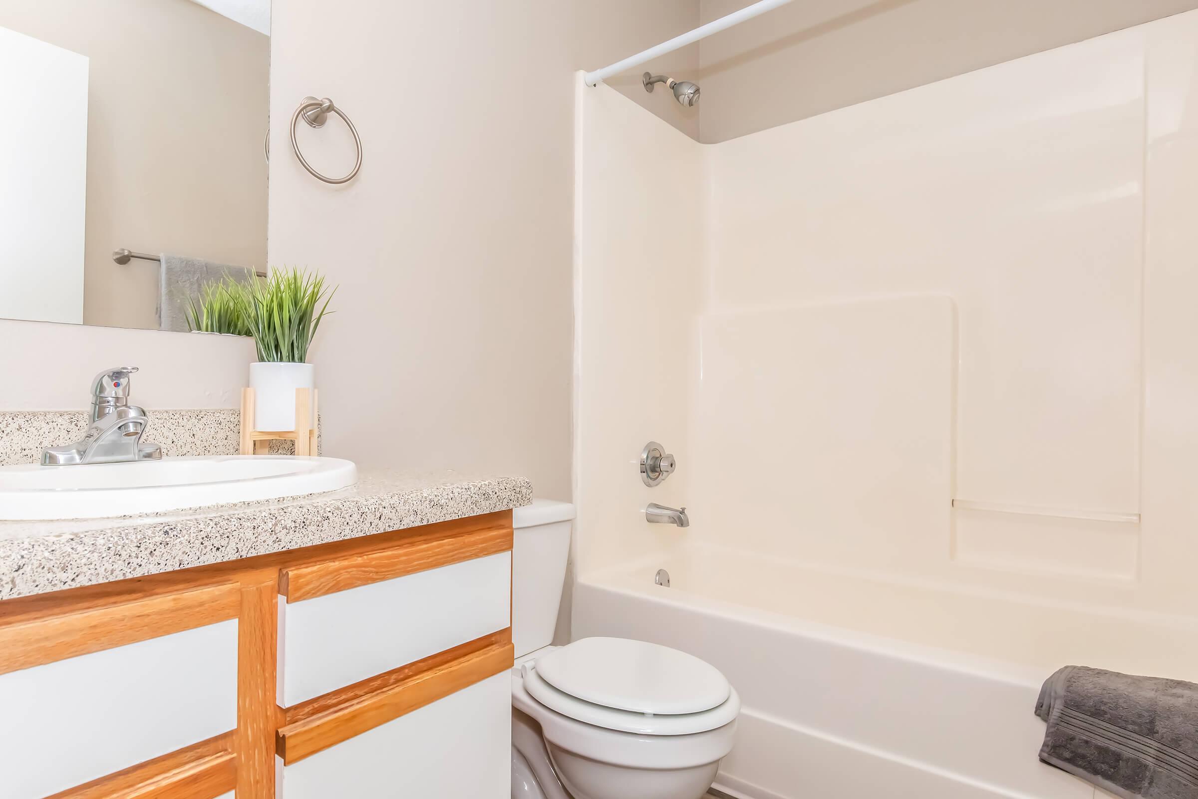 A modern bathroom featuring a beige color scheme. The space includes a white bathtub with a shower curtain, a vanity with a sink, wooden cabinet drawers, a round mirror, and a small plant on the counter. A gray towel is neatly folded on the edge of the tub. The overall look is clean and inviting.