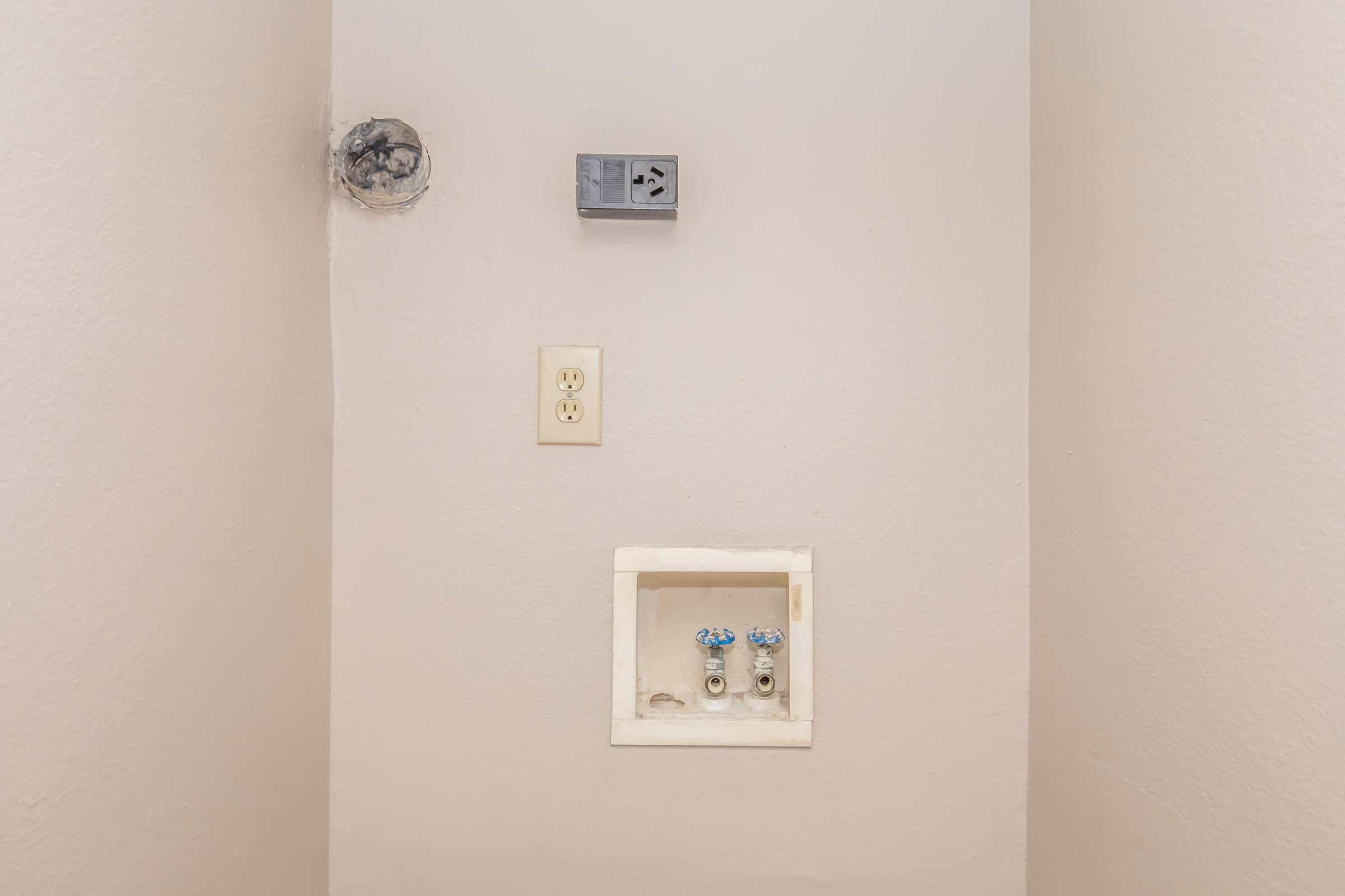 A plain wall featuring two plumbing connections with blue knobs at the bottom, an electrical outlet in the middle, and a small box above. There's a circular vent or hole at the top left corner, and the wall is painted a light color.