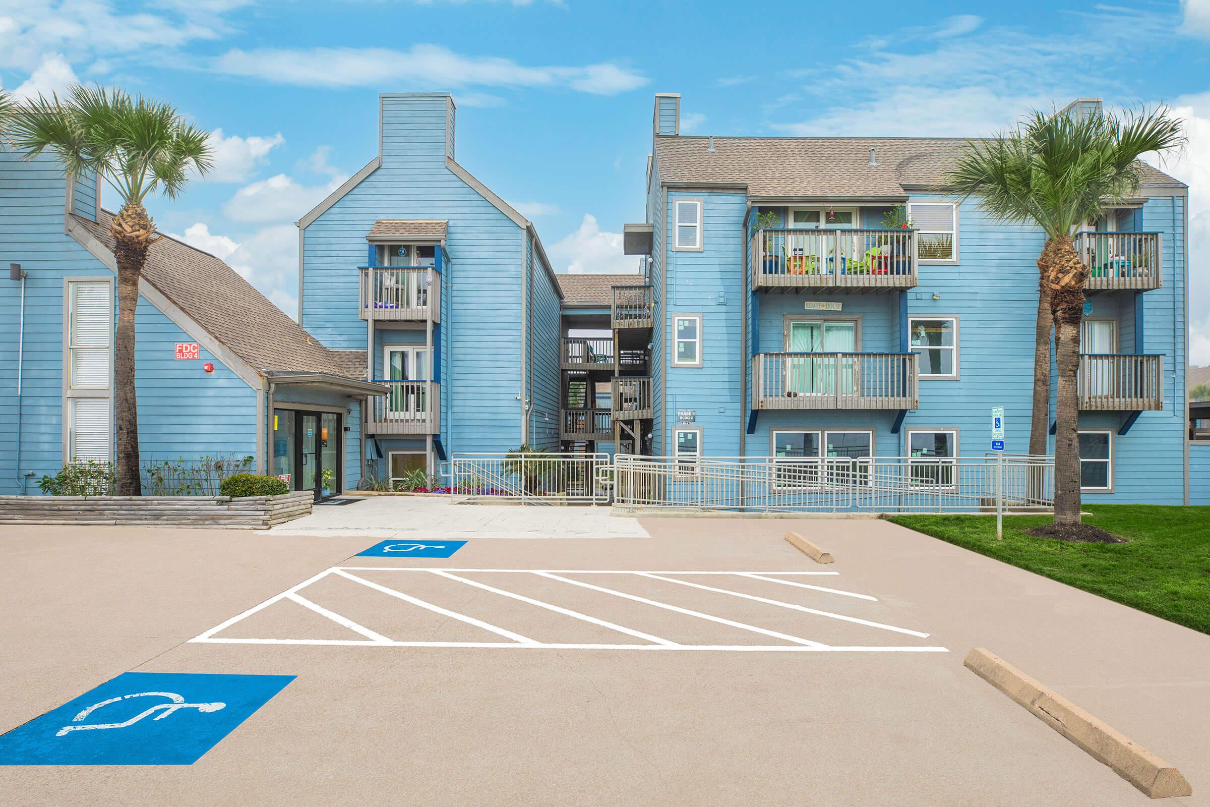 A light blue apartment building with balconies, palm trees in the foreground, and a parking lot featuring designated handicapped spaces. The sky is clear with a few scattered clouds. The building appears well-maintained and inviting.