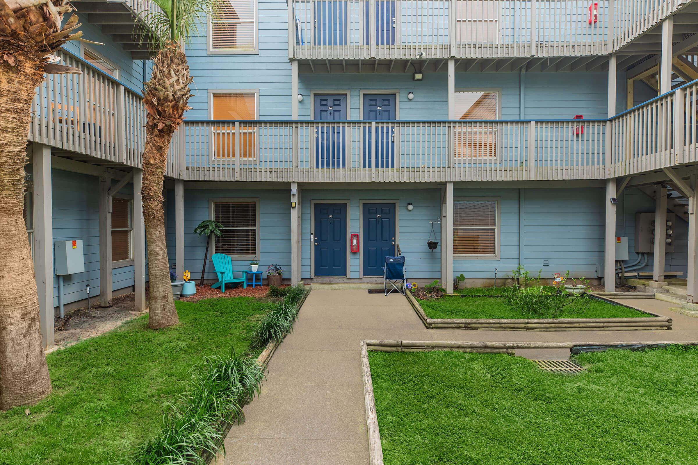A view of a multi-story apartment complex with blue exterior walls. The courtyard features landscaped areas with grass and plants, two palm trees, and a concrete pathway leading to two blue doors. There are also blue chairs and a grill on the patio, creating a cozy outdoor space.