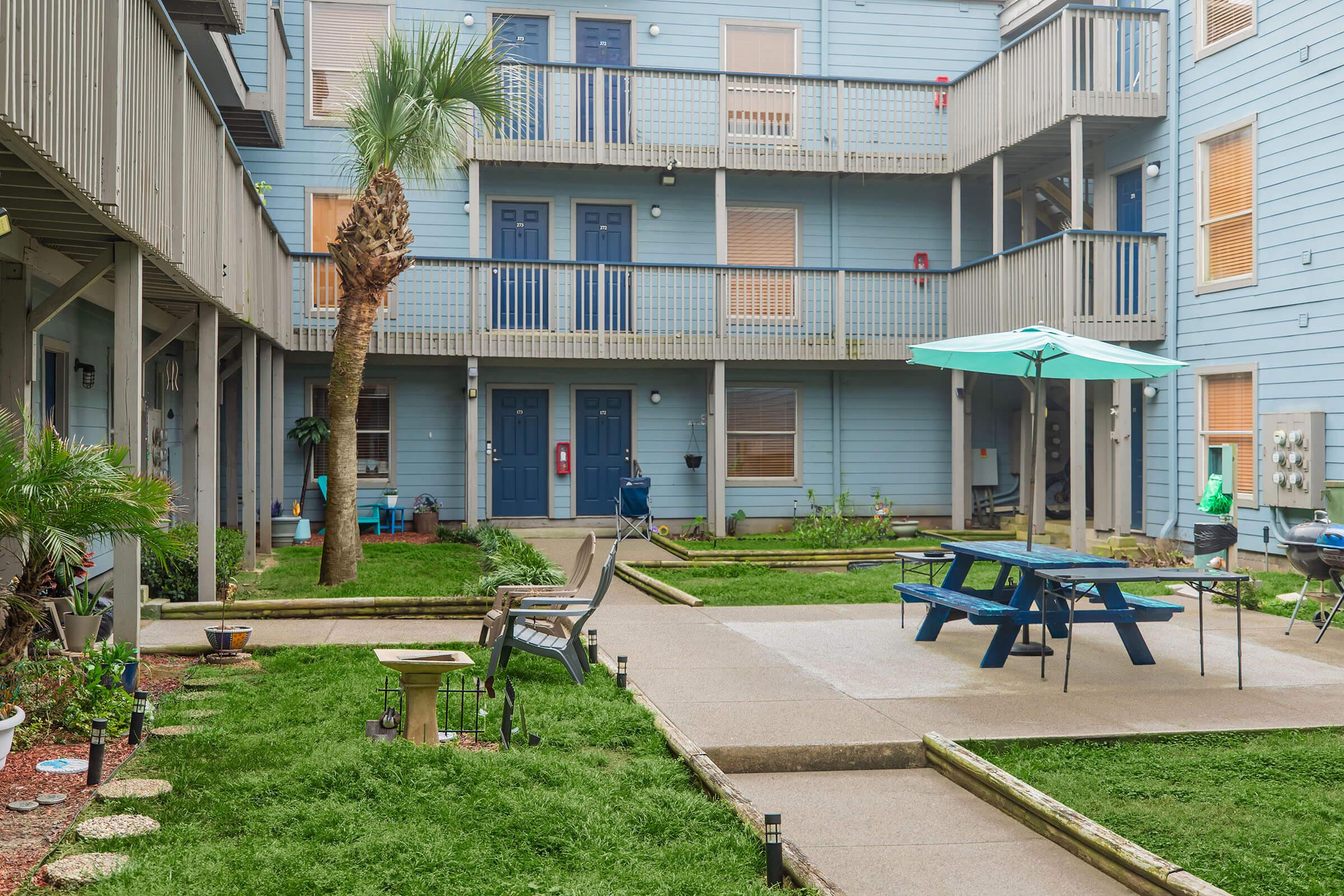 A courtyard of a residential complex featuring a palm tree, picnic table with an umbrella, and landscaped areas. Multiple units with blue siding and balconies are visible in the background, along with garden decorations and pathway lighting. The atmosphere is relaxed and inviting.