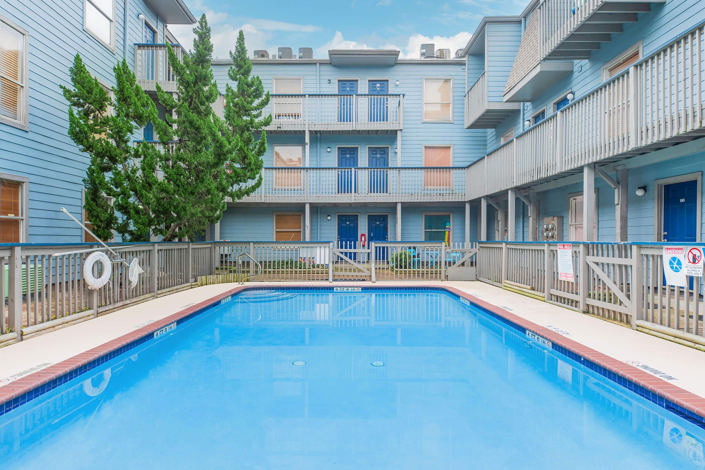 A clear swimming pool surrounded by a wooden deck, with balconies of an apartment building in the background. The building features blue siding and several windows. Lush greenery is visible alongside the pool area.