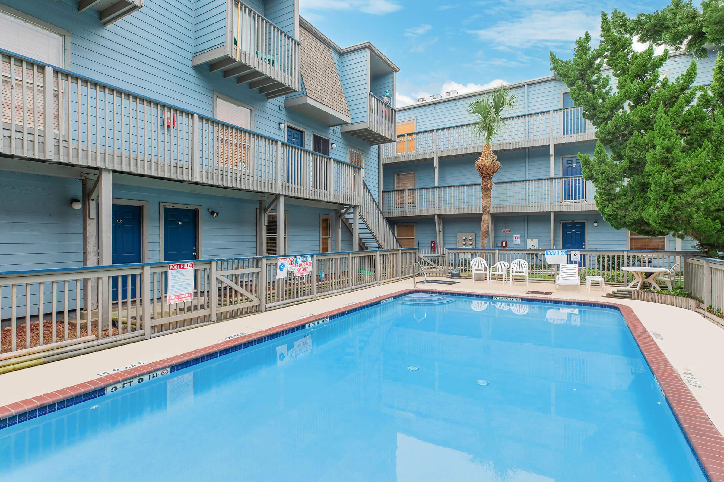 A view of a multi-story building with blue siding surrounding a clear blue swimming pool. The area features outdoor seating and palm trees, with banners and signs visible on the balconies. The sky is partly cloudy, creating a bright and inviting atmosphere.