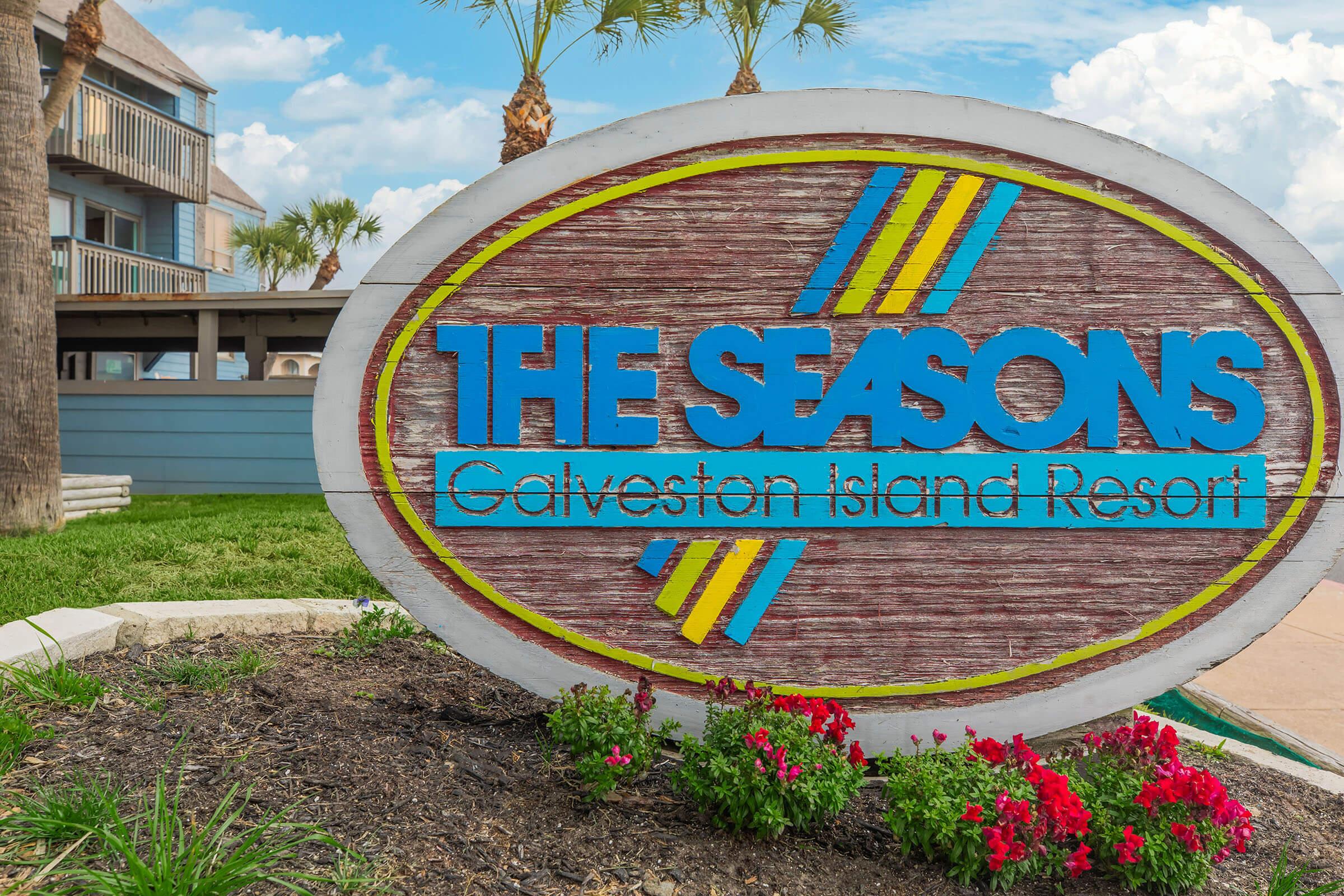 Sign for "The Seasons Galveston Island Resort" featuring blue and yellow lettering on a wooden background, surrounded by greenery and colorful flowers in the foreground, with a blue sky and clouds in the background.