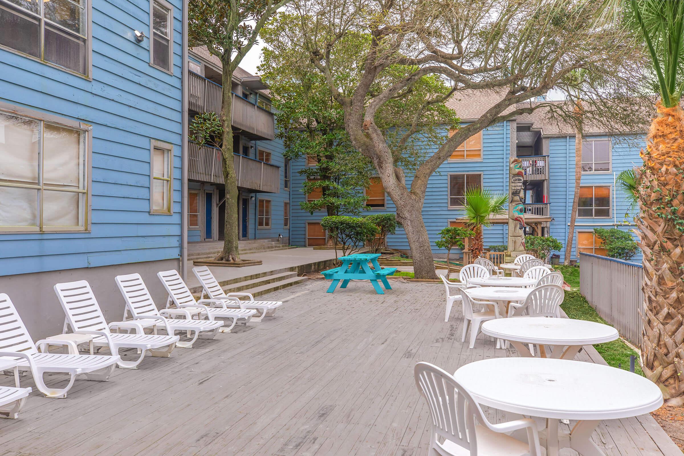 A courtyard with blue wooden buildings surrounding it. The area features white lounge chairs, round tables, and a bright blue picnic table under trees. The setting is peaceful, with greenery and a mix of shaded and sunlit spaces.