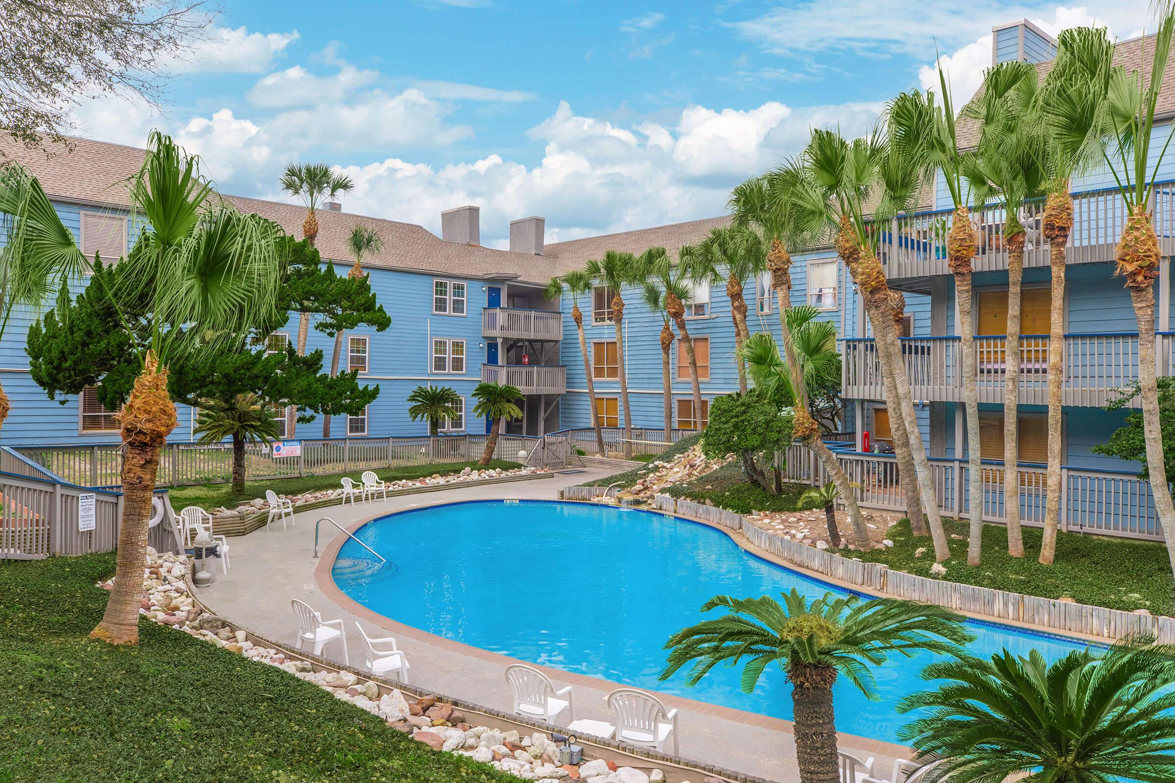 A serene outdoor pool area surrounded by palm trees and colorful apartment buildings. The pool features a clean, blue surface with white lounge chairs nearby. Lush greenery and rocks enhance the tropical atmosphere, while the sky above is bright and partly cloudy.