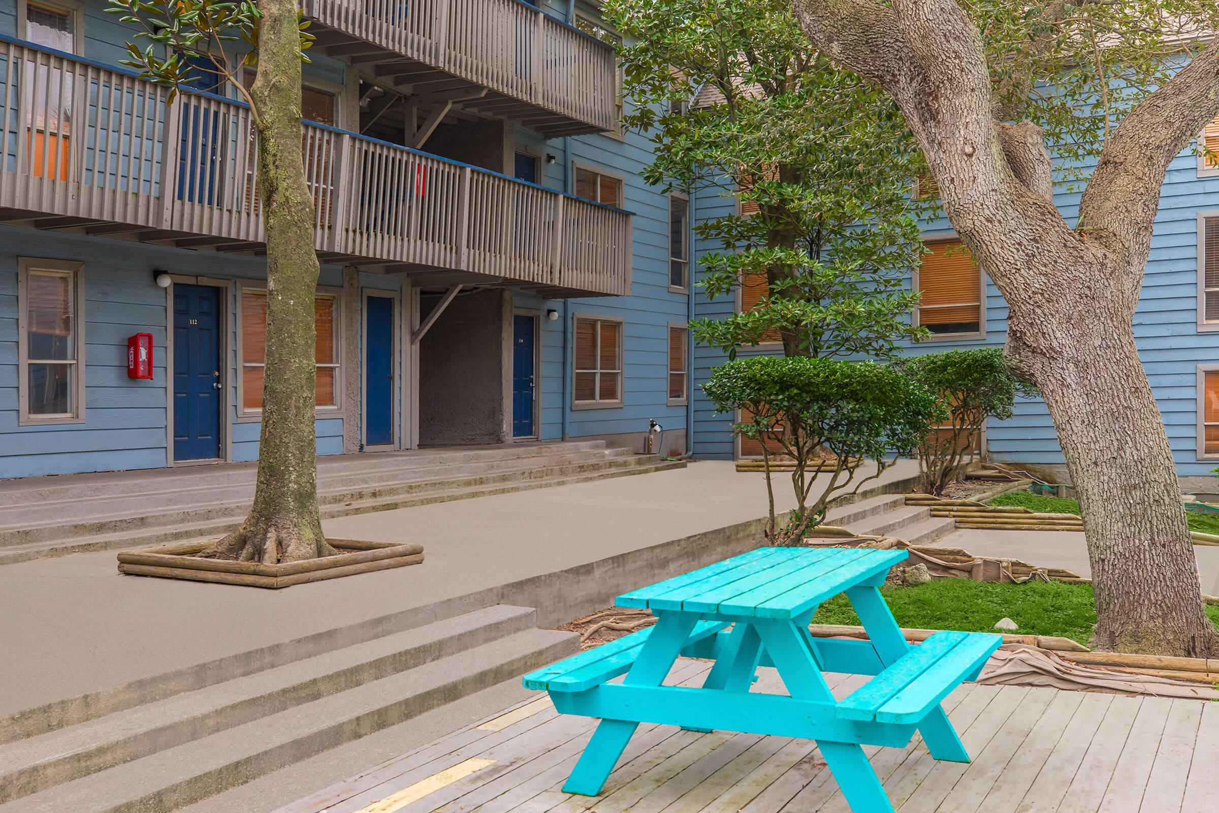 A light blue picnic table sits on a wooden deck in a courtyard surrounded by tree branches and shrubbery, with multiple blue buildings featuring balconies and windows in the background. The setting is calm and inviting, with a peaceful atmosphere.