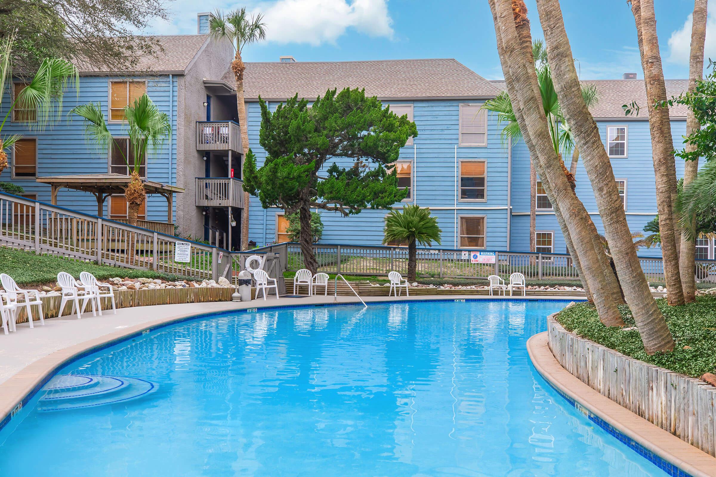 A clear blue swimming pool surrounded by palm trees and lush greenery, with a light blue apartment building in the background. There are white lounge chairs along the poolside and a pathway leading up to the building. The scene is bright and inviting, set in a sunny outdoor setting.