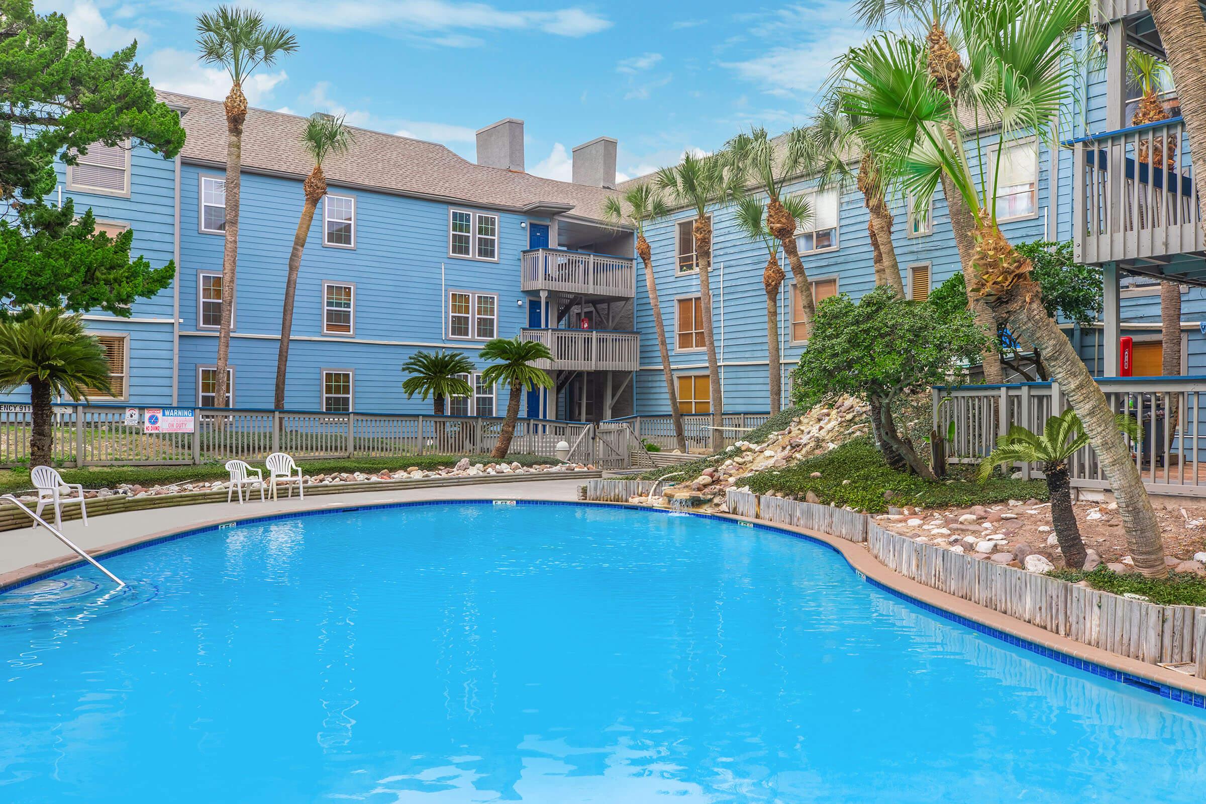 A clear blue swimming pool surrounded by tropical landscaping, with palm trees and a wooden fence. Nearby, a light blue apartment building with multiple balconies can be seen, along with lounge chairs positioned around the pool area. The sky is bright with fluffy clouds.
