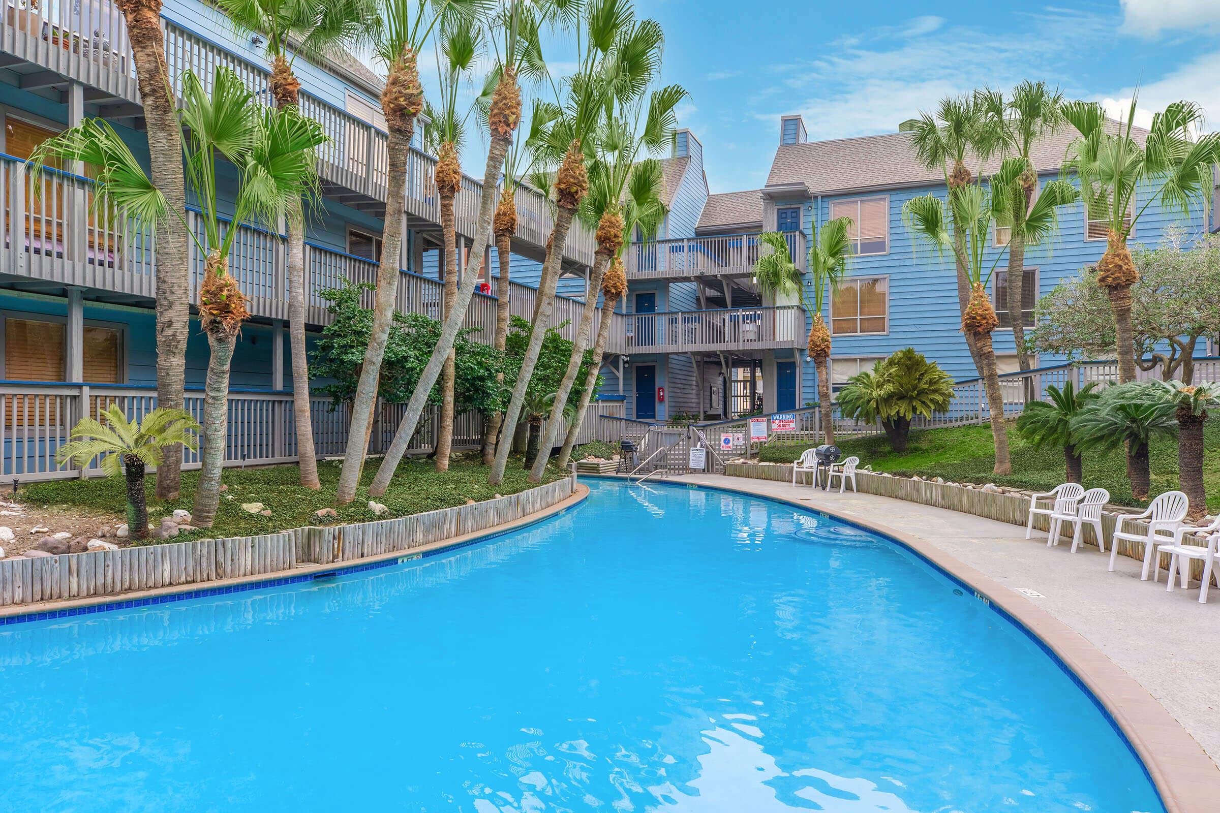 A tranquil pool area surrounded by palm trees and colorful buildings. The pool features a winding shape with lounge chairs placed nearby. The setting includes a lush garden, creating a relaxing atmosphere ideal for leisure and enjoyment. Bright blue skies add to the inviting ambiance.