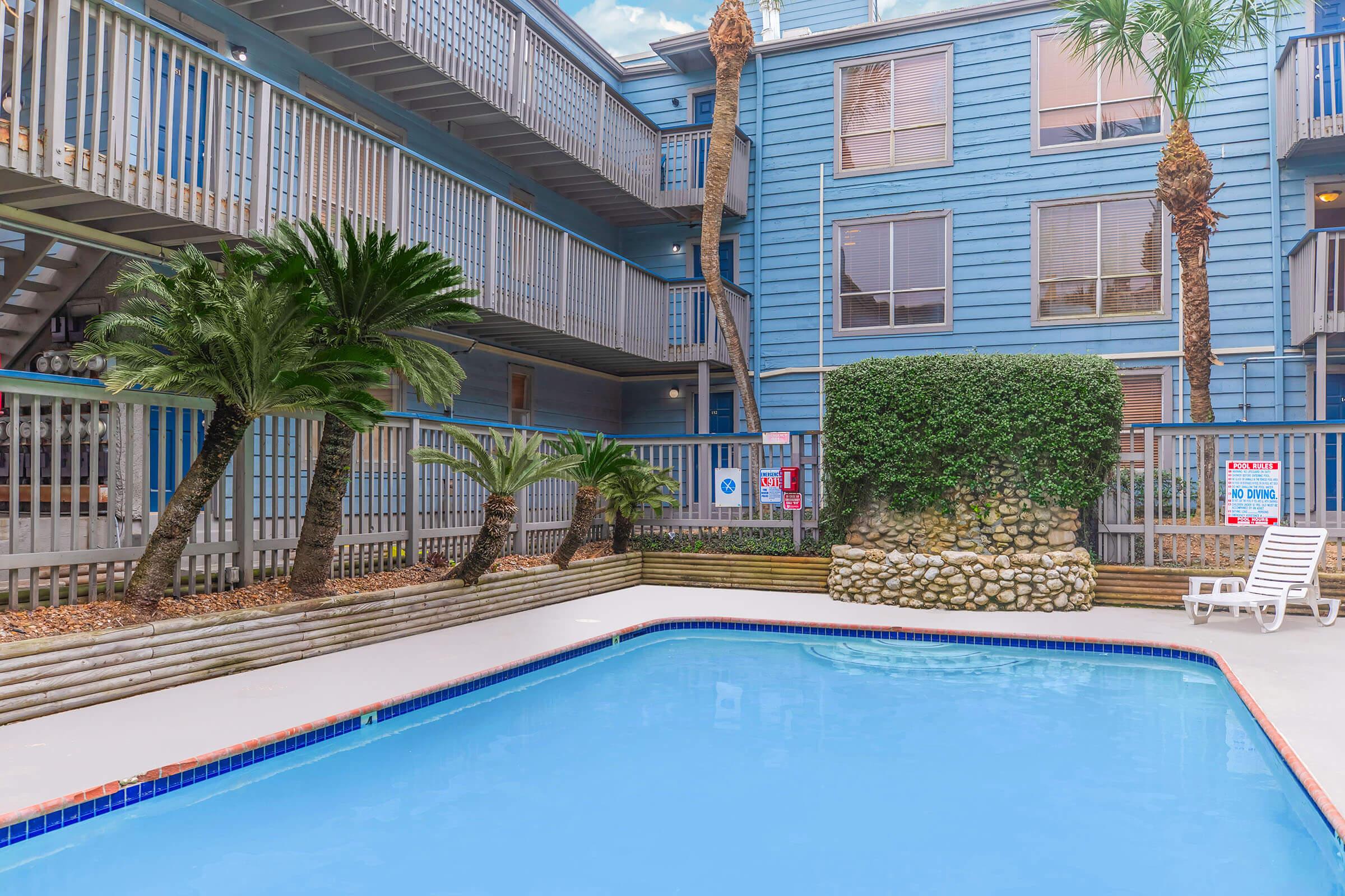 A calm courtyard featuring a swimming pool surrounded by palm trees and a decorative stone wall. The pool area has lounge chairs and is bordered by a blue multi-story building with balconies. Signs and safety features are visible in the background.