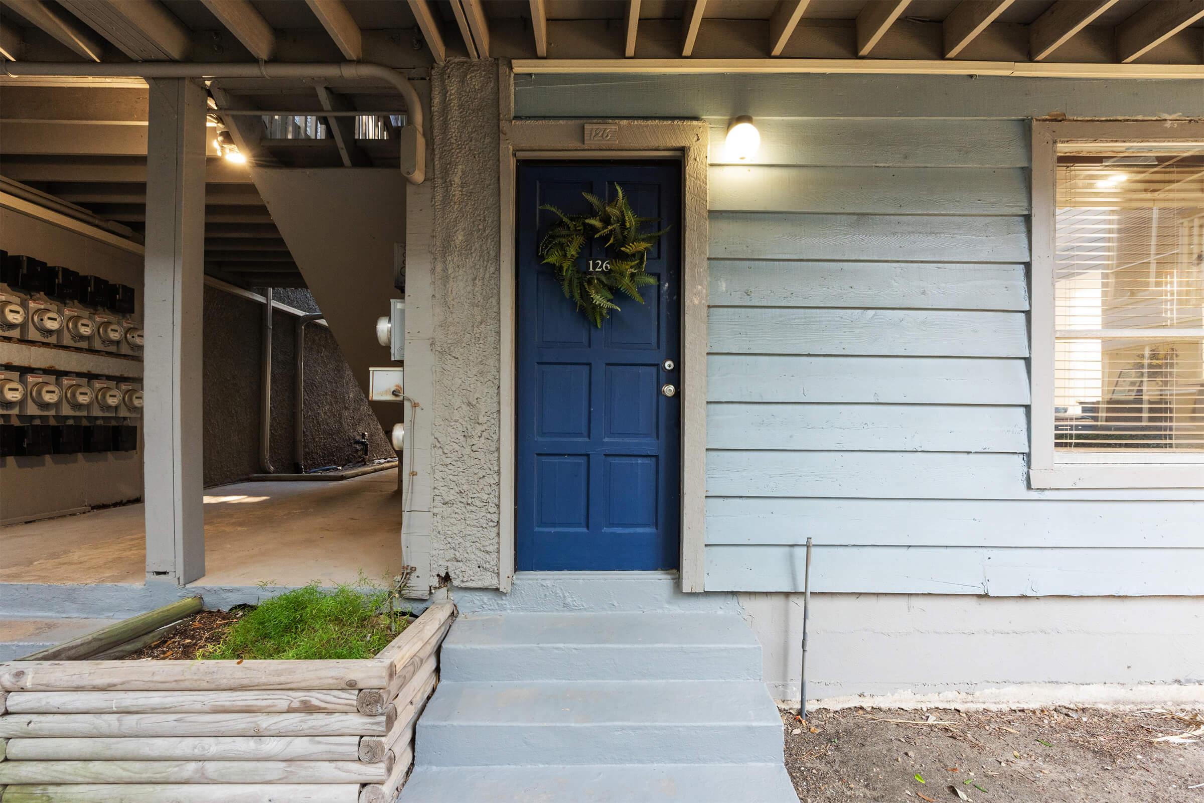 A blue door with a green wreath, set against a light blue wooden wall. The entrance has a small set of concrete steps leading up to the door, with a planter box at the base. Visible in the background are electrical meters mounted on the wall and a partially covered area beneath the building.