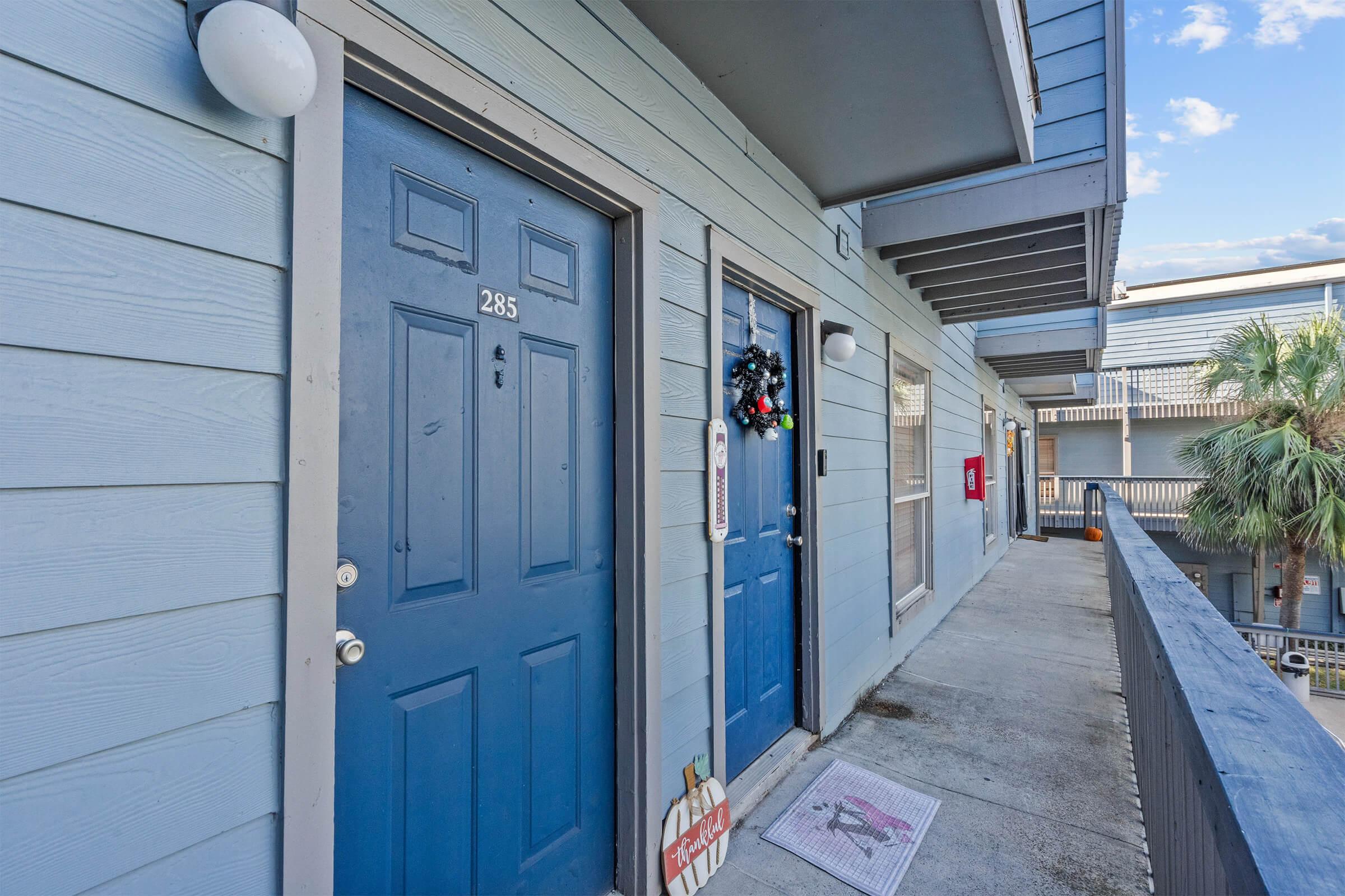 Two blue doors labeled "285" and another door, located along a light blue exterior corridor. A decorative wreath is hanging on the door beside the number 285. The area is bright and has a clear blue sky in the background, with some greenery visible, suggesting a pleasant environment.