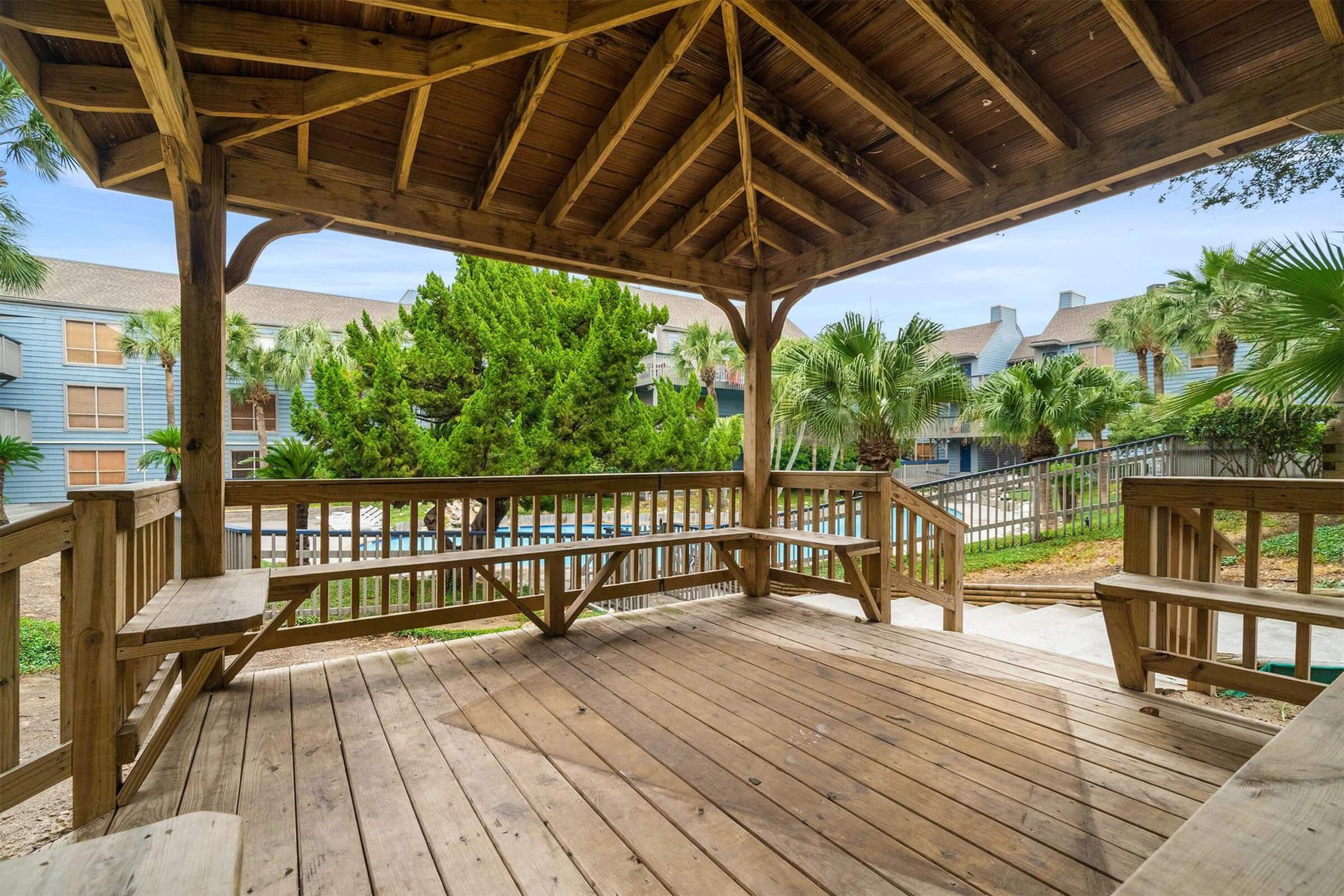 A wooden gazebo with a spacious deck and seating, surrounded by green palm trees and a glimpse of a swimming pool in the background. The structure has a steep roof and open sides, inviting relaxation in a tropical setting.