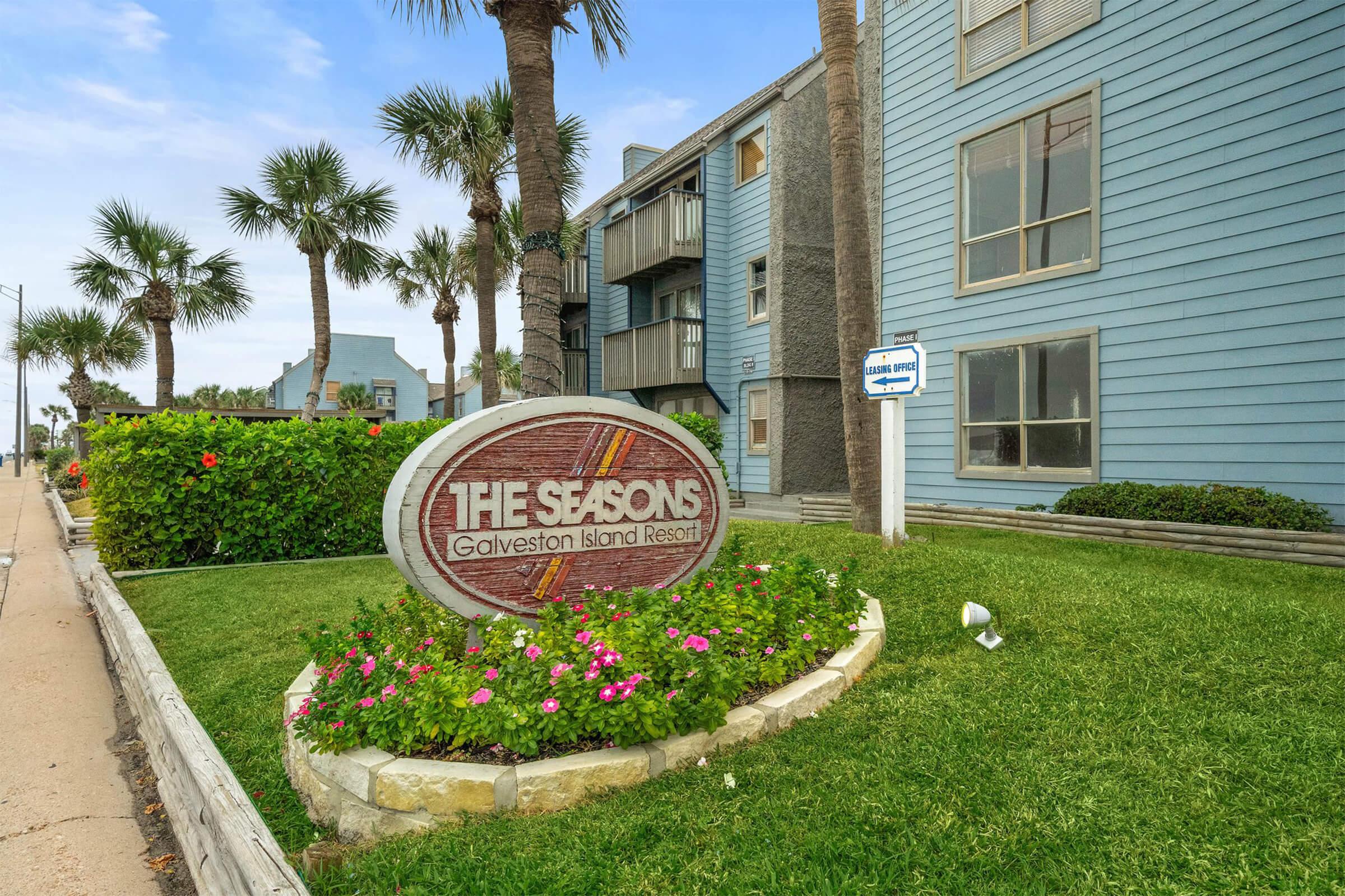 Sign for "The Seasons Galveston Island Resort" in front of a multi-story building. The area features palm trees and well-maintained landscaping with flowers and shrubs, set against a clear sky. A sidewalk runs alongside the property, indicating proximity to the street.