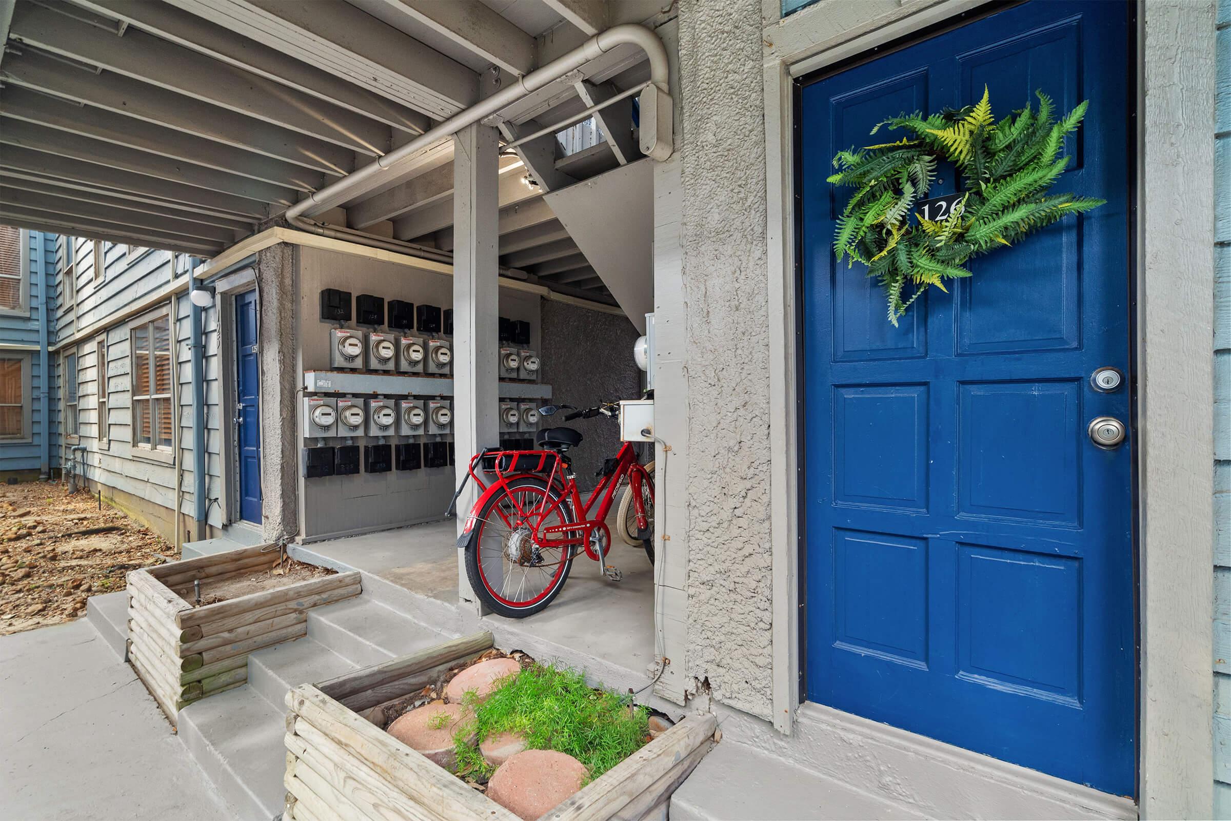 Entrance to an apartment building featuring a blue door adorned with a green wreath. To the left, a red bike is parked, and there are mailboxes mounted on the wall. Below the entrance, there are planters with green plants and decorative stones. The surroundings include a wooden overhang and multiple stories of the building.