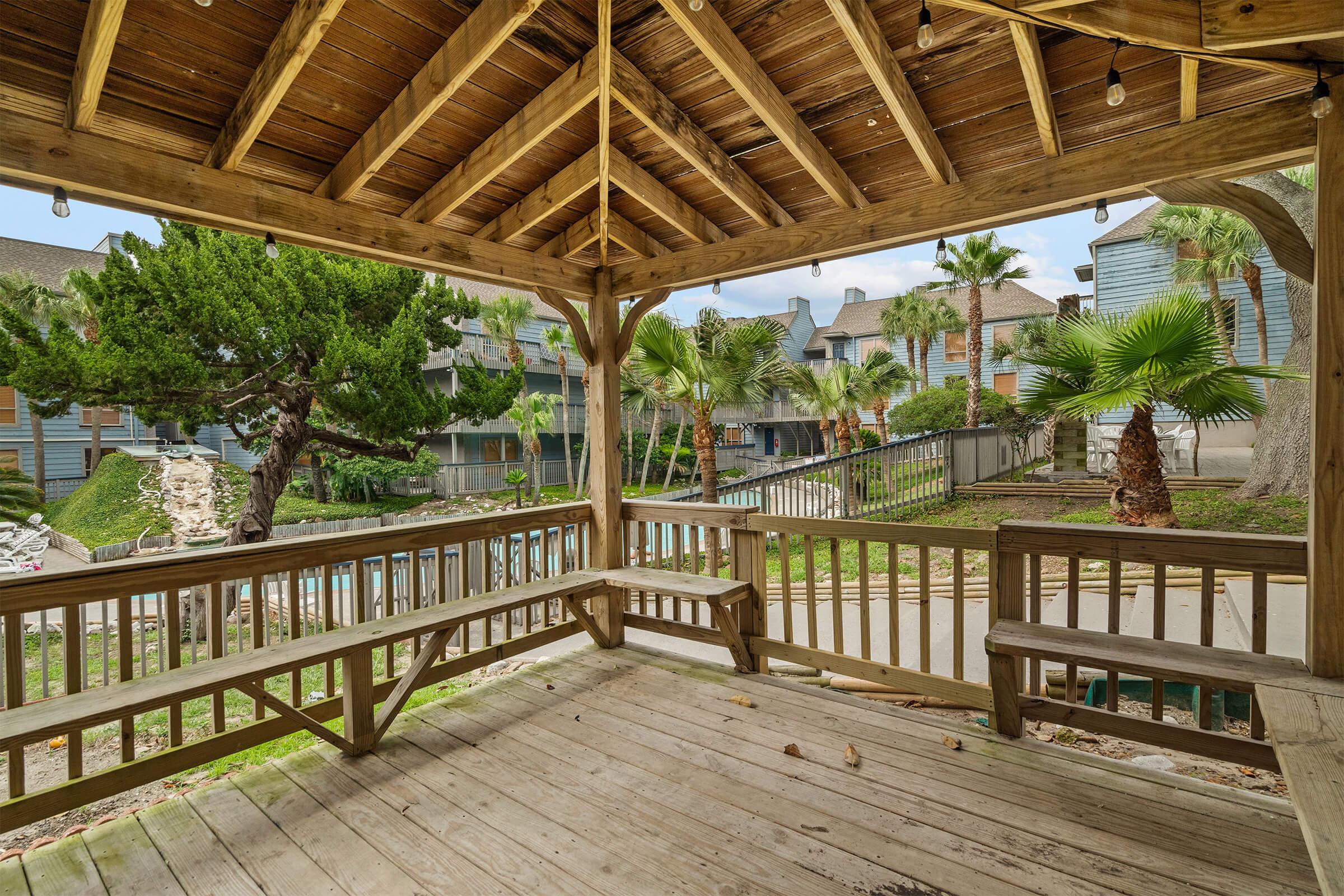 A wooden gazebo with a sloped roof, surrounded by palm trees and a landscaped area. There are benches along the sides, and the view features multi-story buildings in the background, adding to the serene outdoor setting. Soft lighting strings hang from the roof beams, enhancing the atmosphere.