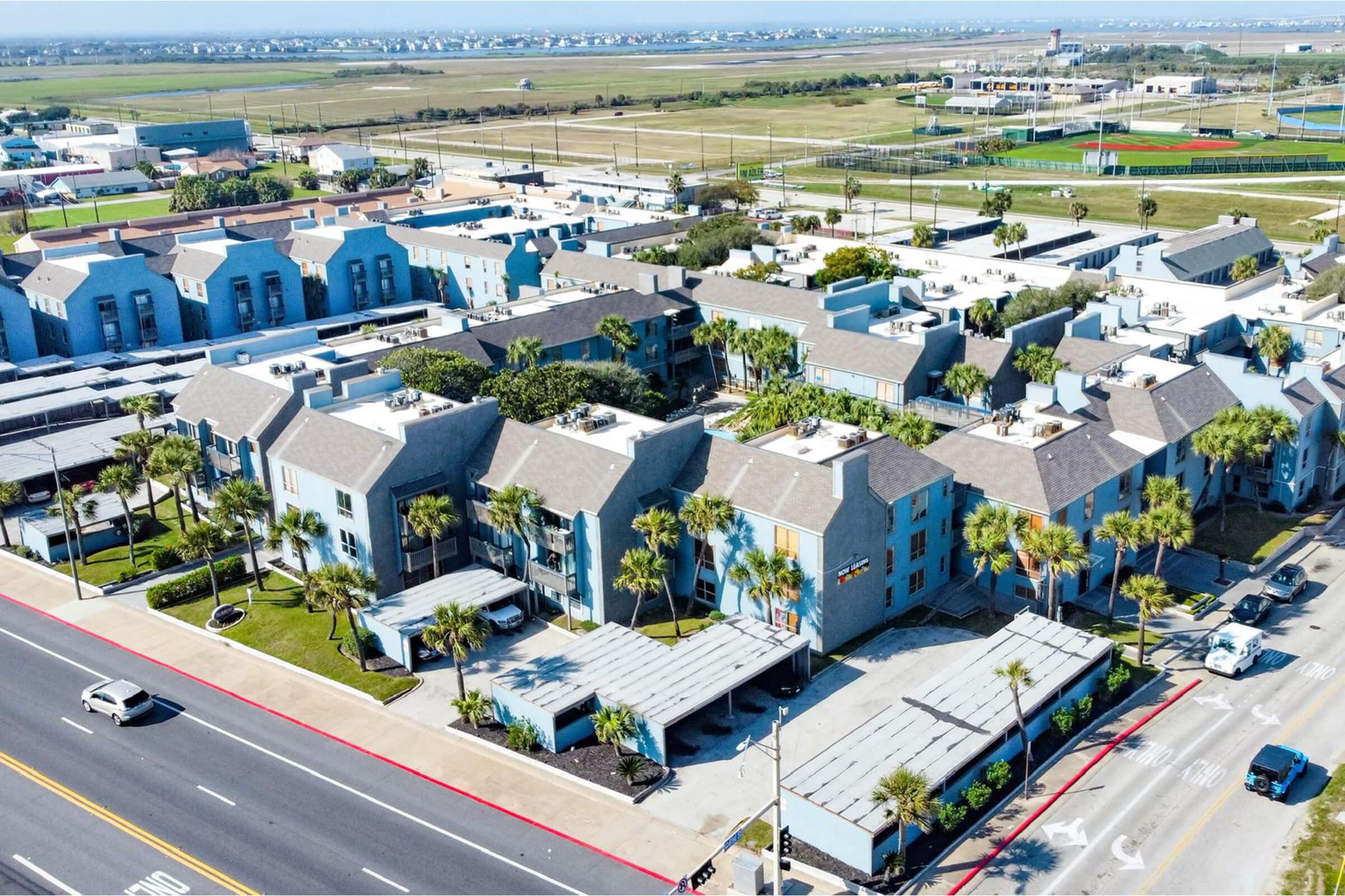 Aerial view of an apartment complex with multiple blue buildings, palm trees, and parking spaces. In the background, a flat landscape stretches out, with a glimpse of a field and distant buildings under a clear blue sky. A road runs along the foreground.