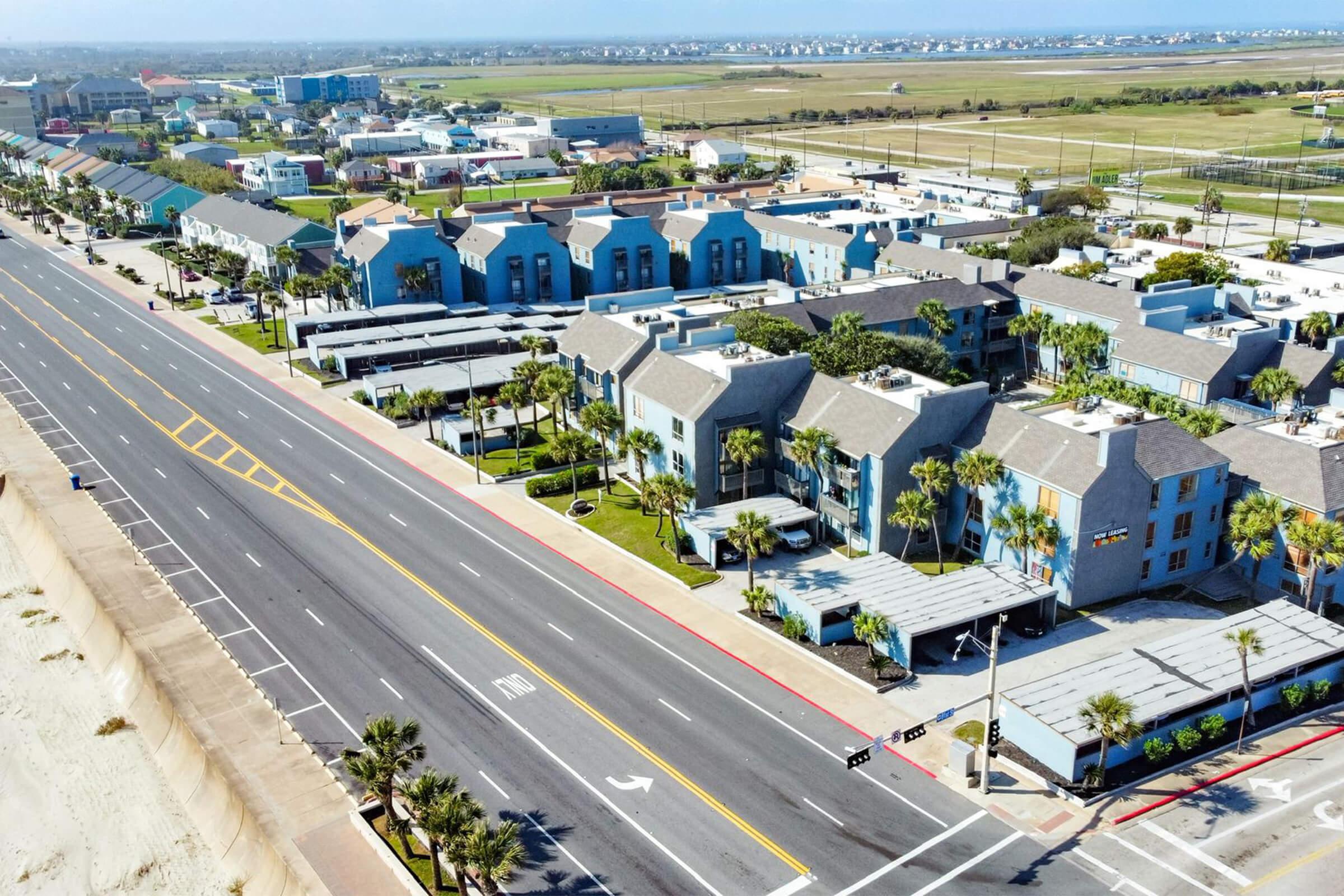 Aerial view of a coastal area featuring hotels and buildings with blue exteriors, palm trees, and a wide road. In the background, there are fields and distant buildings with a clear blue sky.