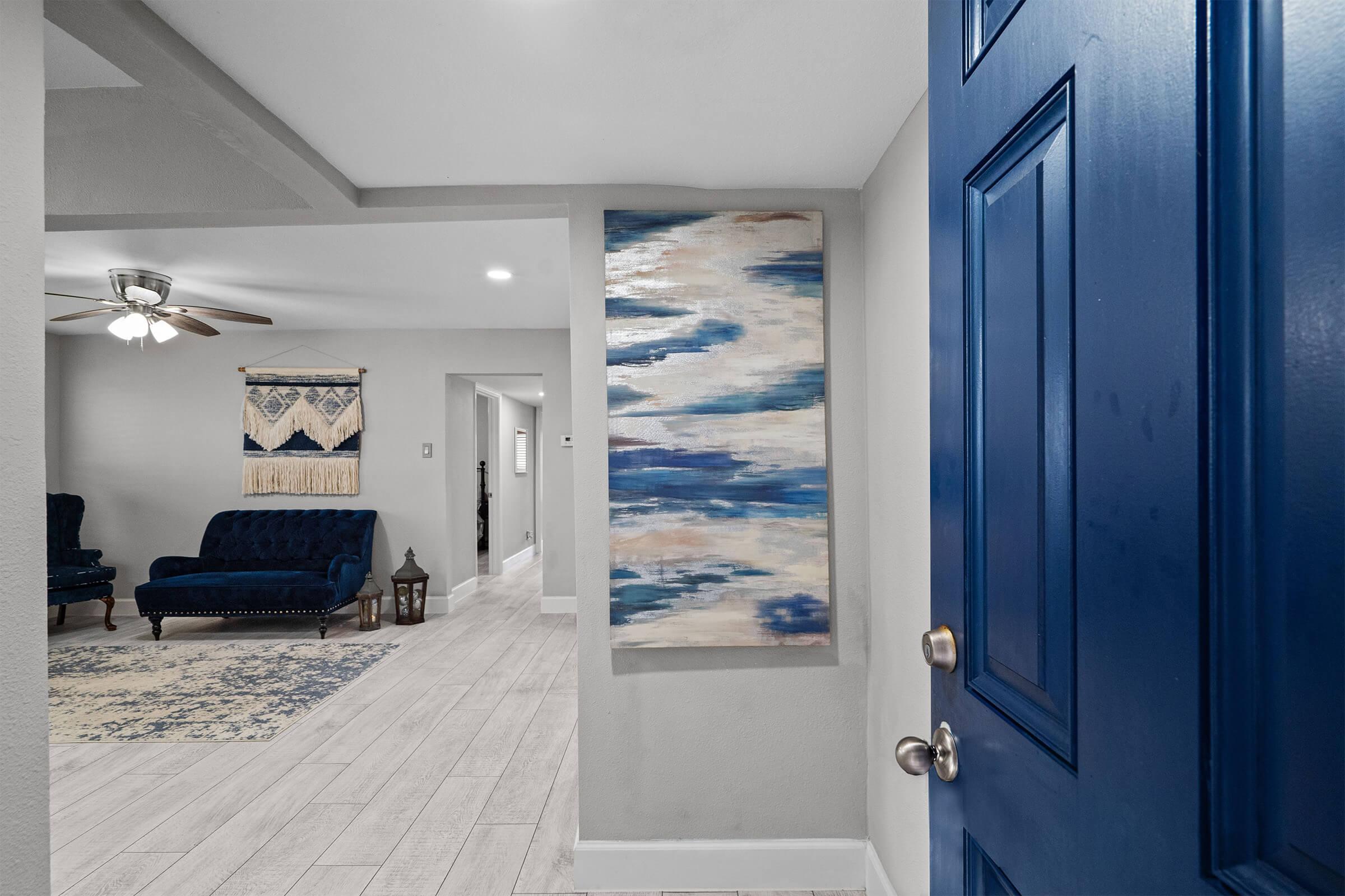 A stylish entryway featuring a vibrant blue front door, a large abstract painting with shades of blue on the wall, and a cozy seating area with dark blue sofas. The space is well-lit with modern decor, showcasing light-colored flooring and a decorative rug.