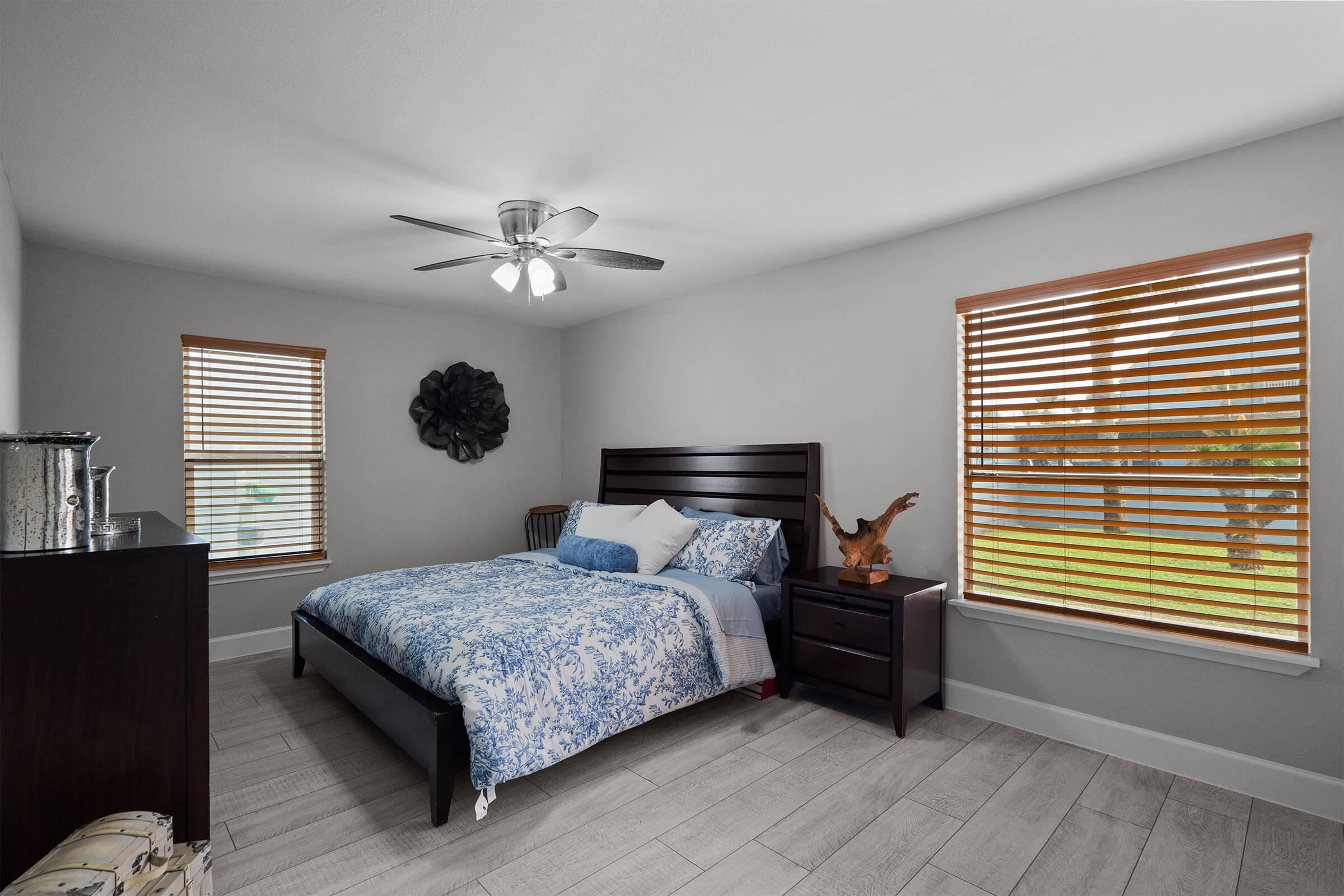 A cozy bedroom featuring a queen-sized bed with a blue and white floral comforter, two wooden nightstands, and a ceiling fan. Large windows with wooden blinds allow natural light and showcase greenery outside. The room has light gray walls and wooden flooring, creating a warm and inviting atmosphere.