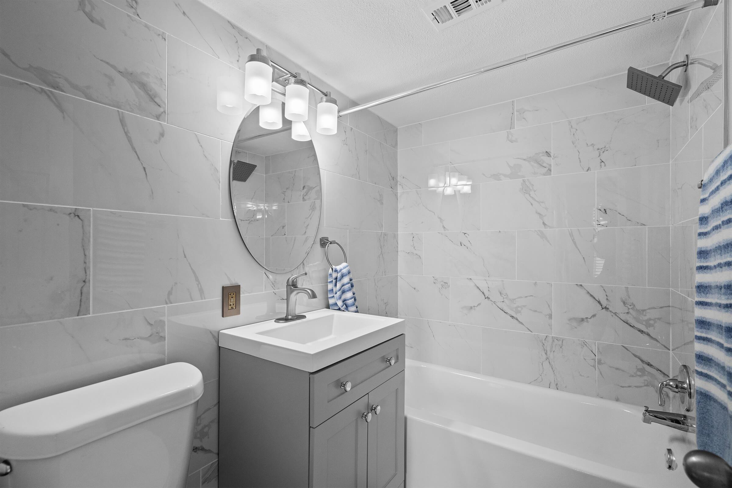 Modern bathroom featuring a light marble tile design, a circular mirror above a white countertop vanity, a silver faucet, and a bathtub. Bright lighting from above enhances the space, while a blue and white striped towel hangs nearby. A toilet is also present in the design.