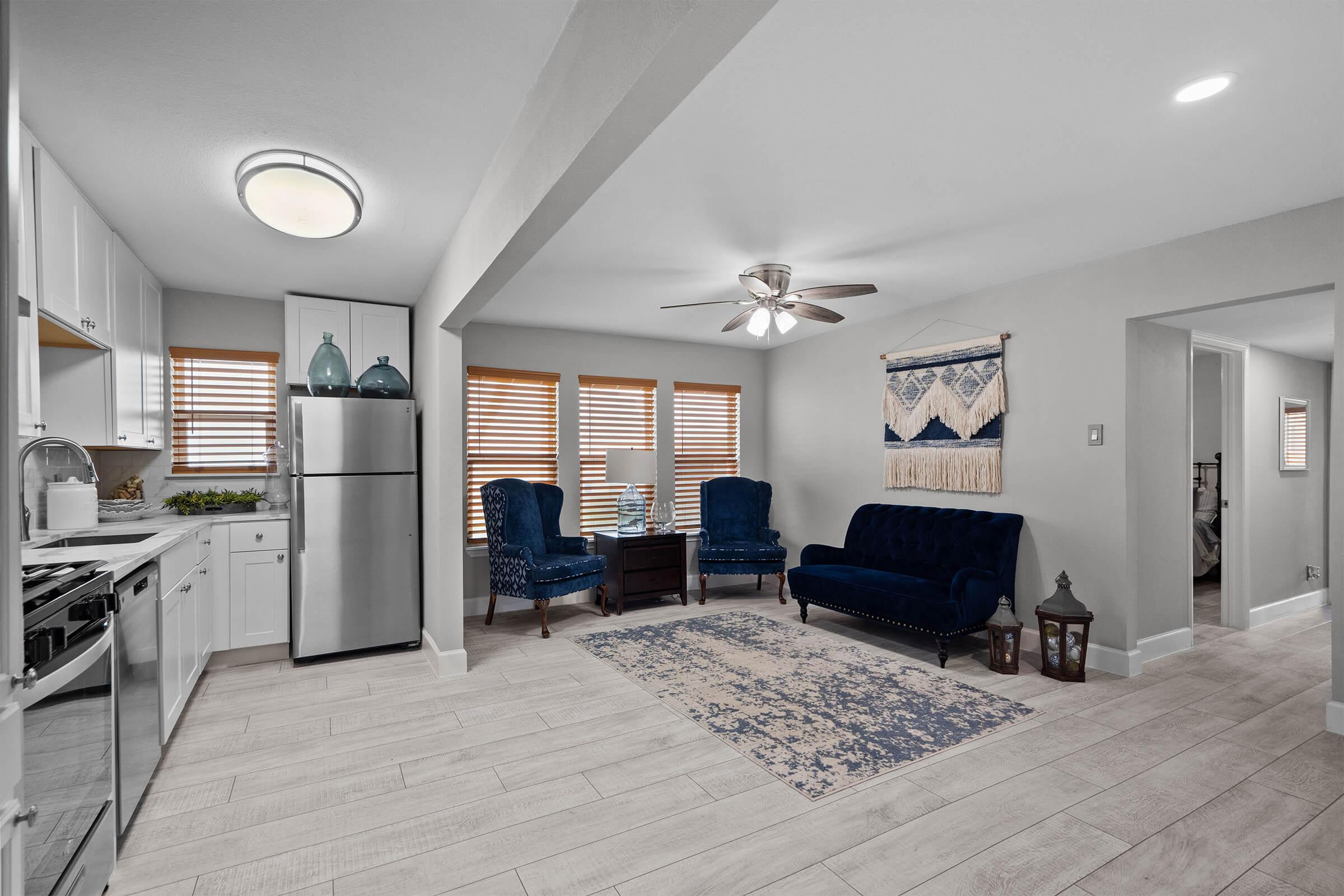 A modern living space featuring a kitchen with stainless steel appliances, a cozy seating area with blue velvet chairs and a matching sofa, a decorative rug, and natural light coming through blinds. The decor includes a wall hanging and a lantern, creating a welcoming atmosphere.