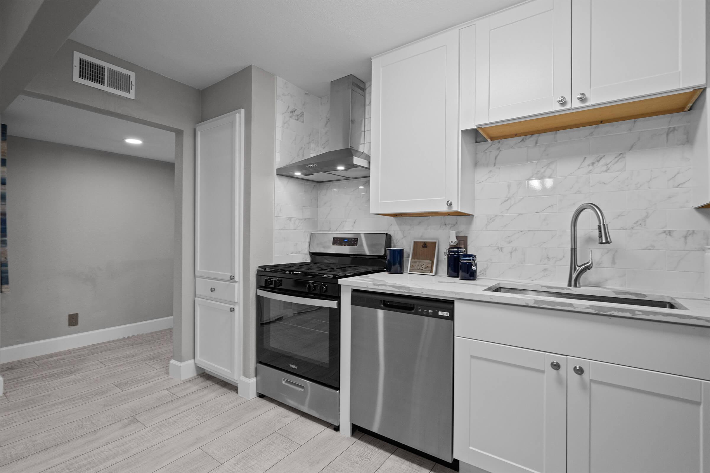 Modern kitchen featuring white cabinetry, marble backsplash, and stainless steel appliances. The gas range is adjacent to a dishwasher, with a sink and modern faucet on the right. Neutral-toned walls and wood-look tile flooring complete the space, creating a clean and inviting atmosphere.