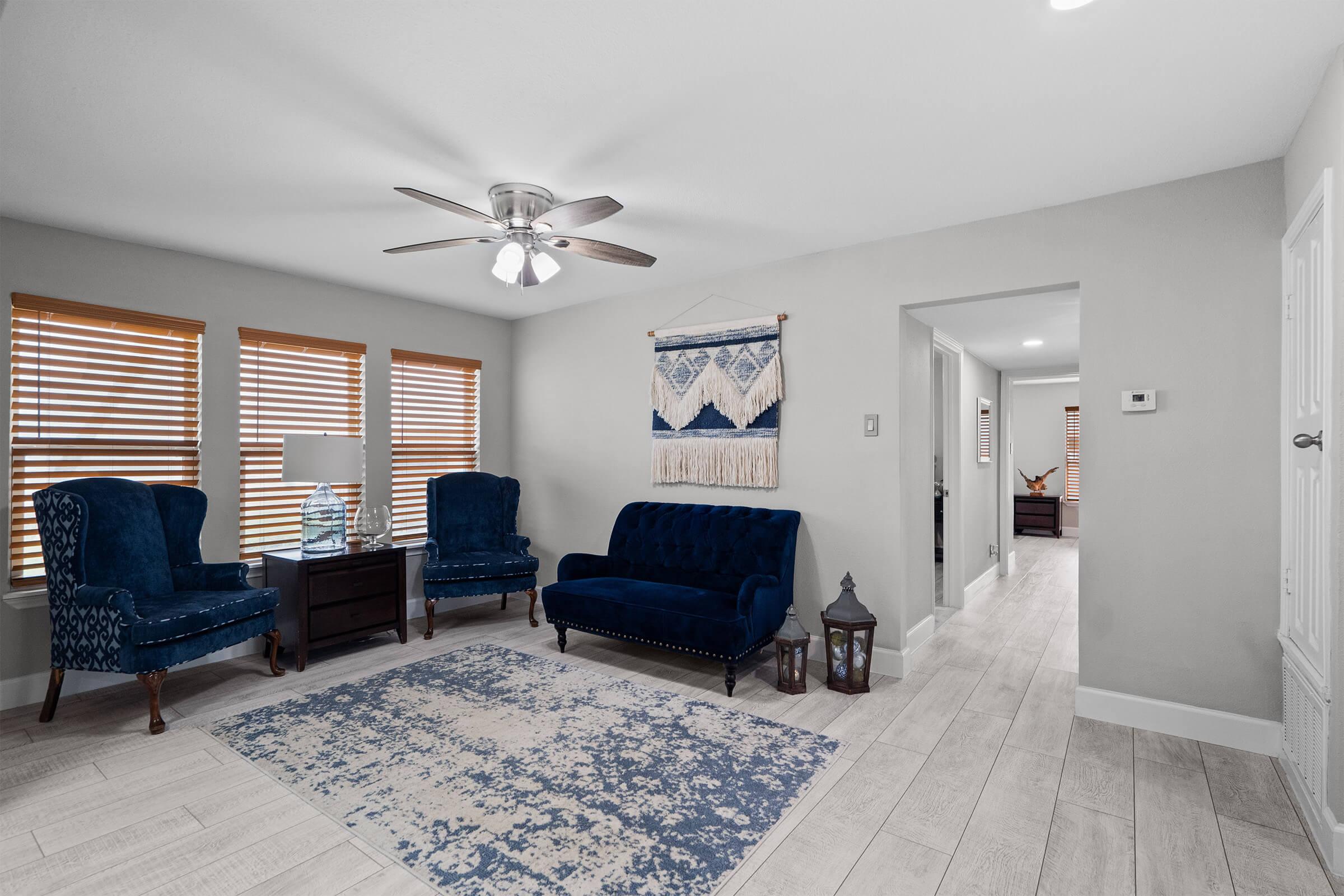 A cozy living room featuring two blue upholstered armchairs and a matching blue sofa. The space has wooden blinds on the windows, a decorative wall hanging, and a light-colored floor. A ceiling fan and a small side table with a lamp are included, creating a welcoming atmosphere.