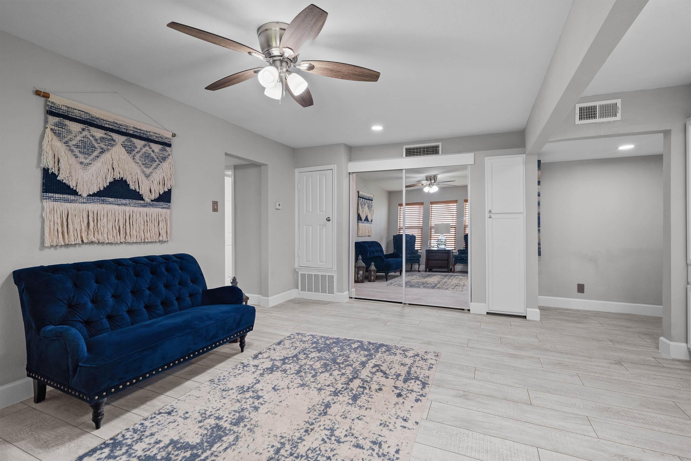 A modern living room featuring a blue tufted sofa, a light-colored area rug, and a ceiling fan. The walls are painted in a neutral shade, and there are mirrored sliding doors on one side. Decorative wall art hangs above the sofa, and the space is well-lit by natural light.