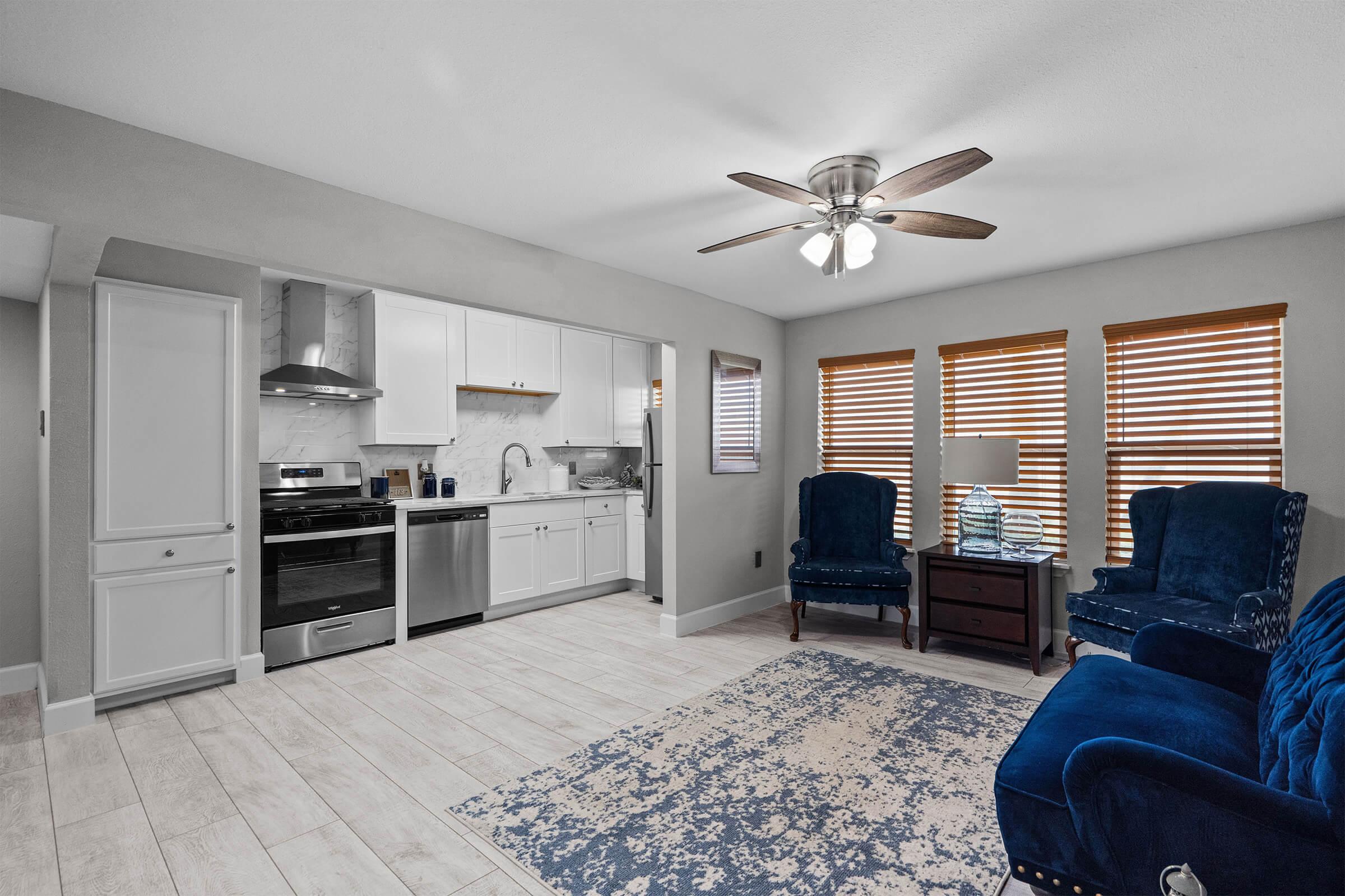 Cozy living space featuring two blue upholstered chairs, a wooden side table, and a patterned area rug. The kitchen area includes modern appliances, white cabinets, and marble countertops. Natural light streams through wooden blinds on the windows, enhancing the inviting atmosphere.
