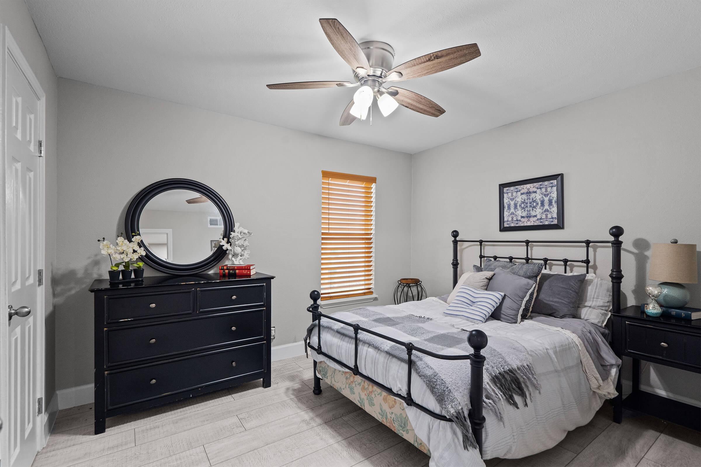 A cozy bedroom featuring a metal bed frame with gray and white bedding, a round mirror on the wall, a dark wood dresser, a bedside table with a lamp, and a window with wooden blinds. The room has light-colored walls and wooden flooring, creating a warm and inviting atmosphere.