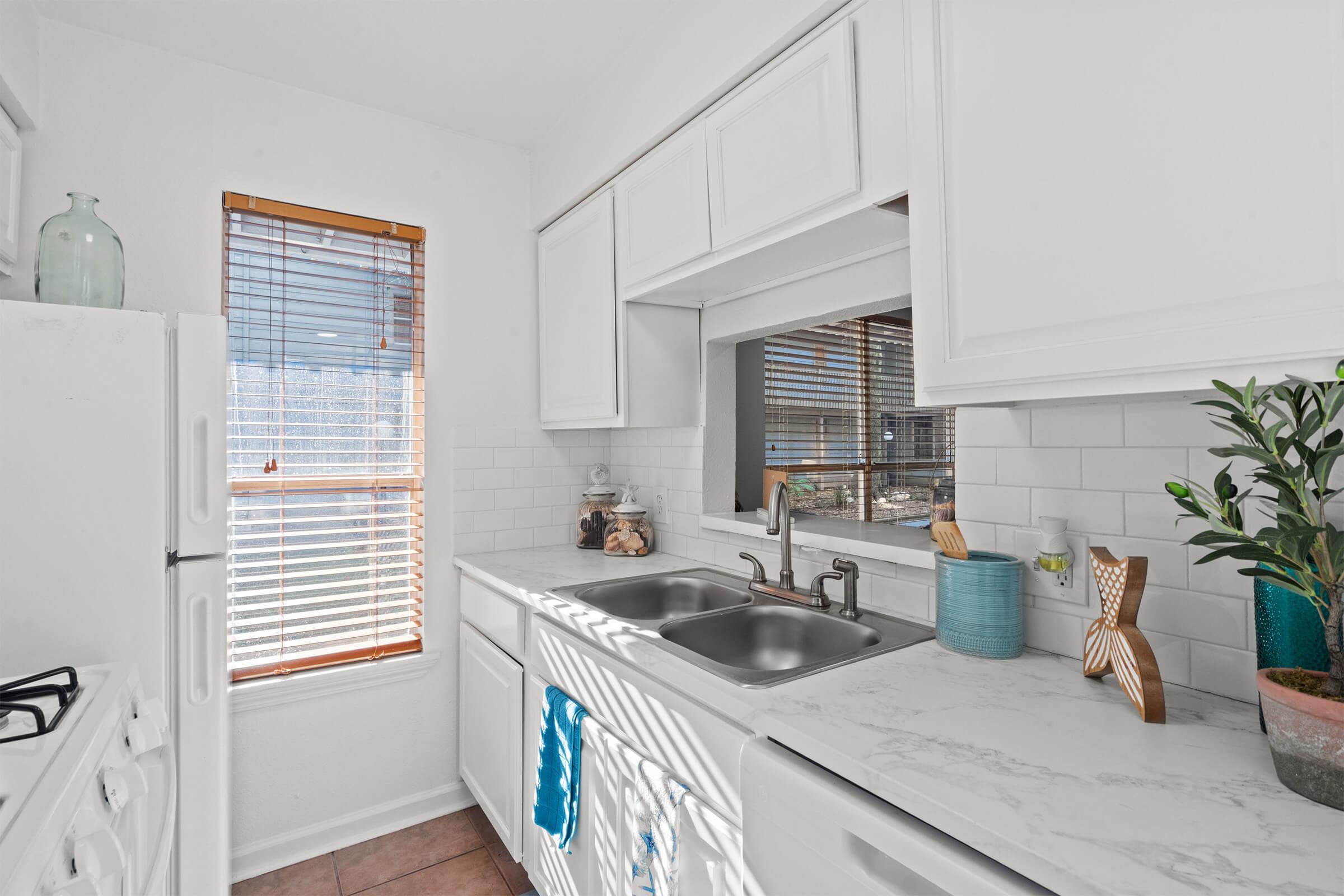 A bright, modern kitchen featuring white cabinetry and a marble countertop. There are double sinks with a faucet, a window with wooden blinds letting in natural light, and a small potted plant nearby. Decorative items, including a teal container and a wooden fish, are placed on the countertop, adding charm to the space.