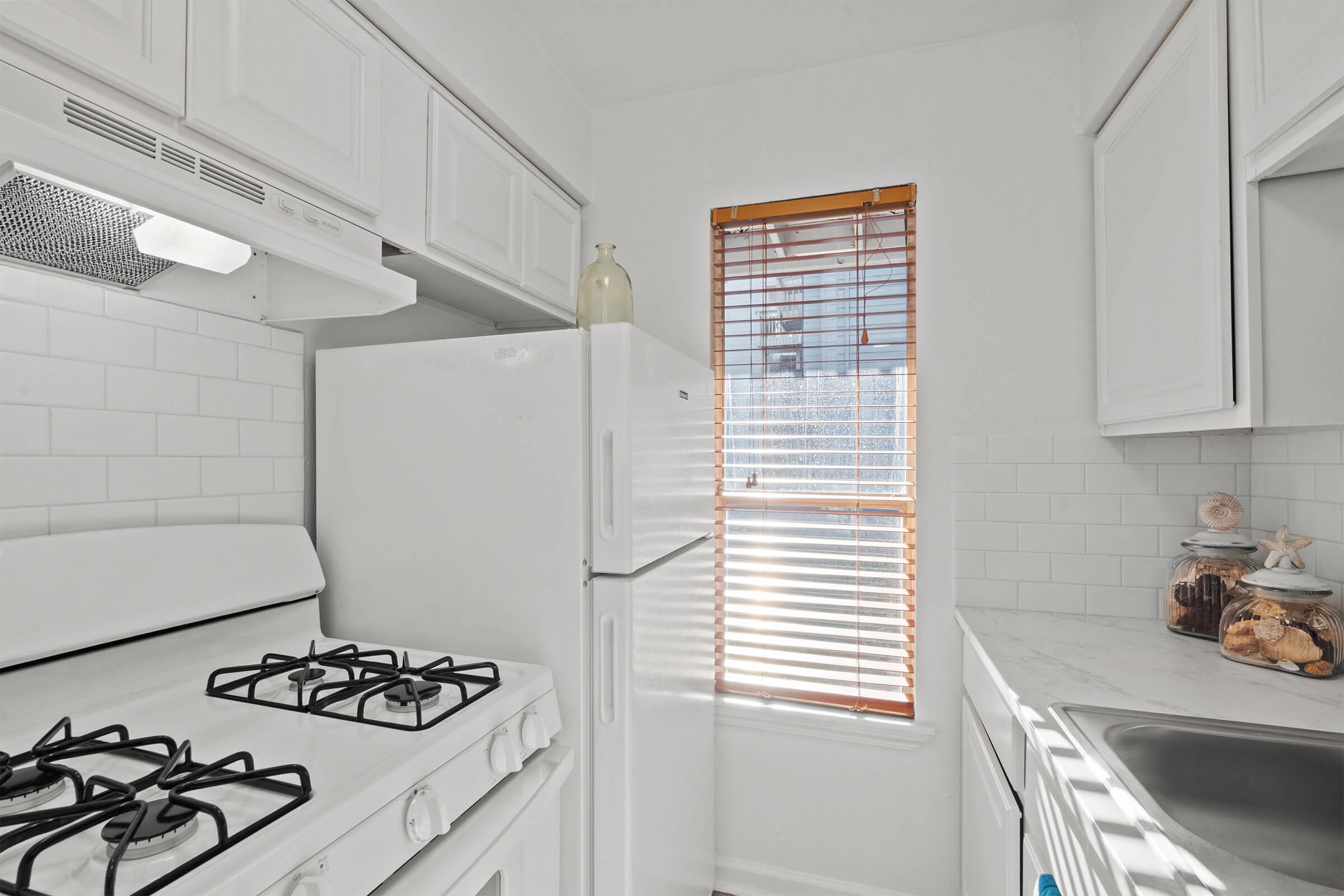 A bright and modern kitchen featuring a white gas stove, refrigerator, white cabinetry, and a marble countertop. Natural light streams in through a partially opened window with wooden blinds, illuminating the space. Decorative items are placed on the counter, adding a cozy touch.