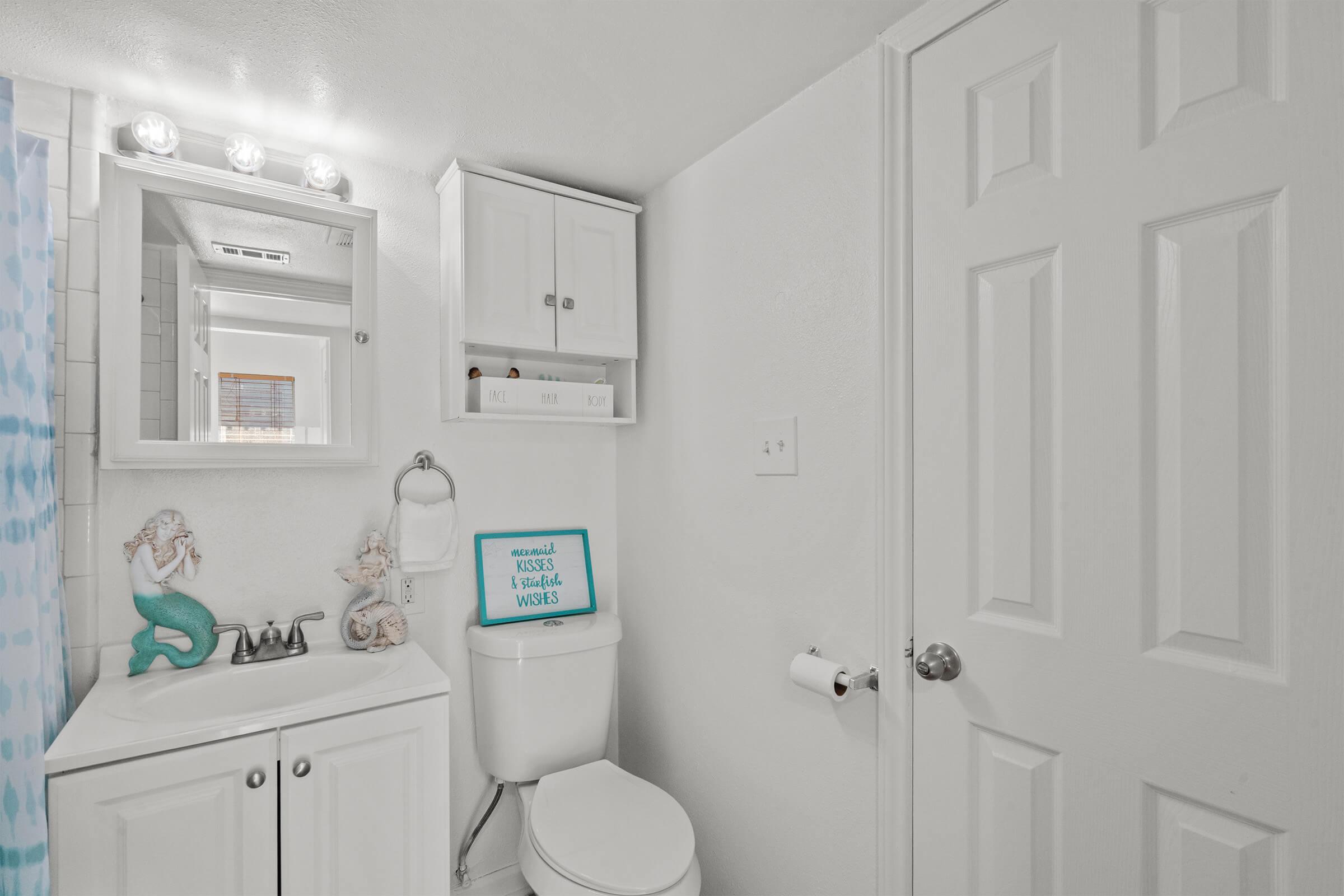A bright, clean bathroom featuring a white toilet, sink with storage underneath, and a lighted mirror. A decorative mermaid figurine is on the sink, and a sign on a shelf reads "Mermaids wish you well." The walls are painted white, and there's a towel rack with a roll of toilet paper.