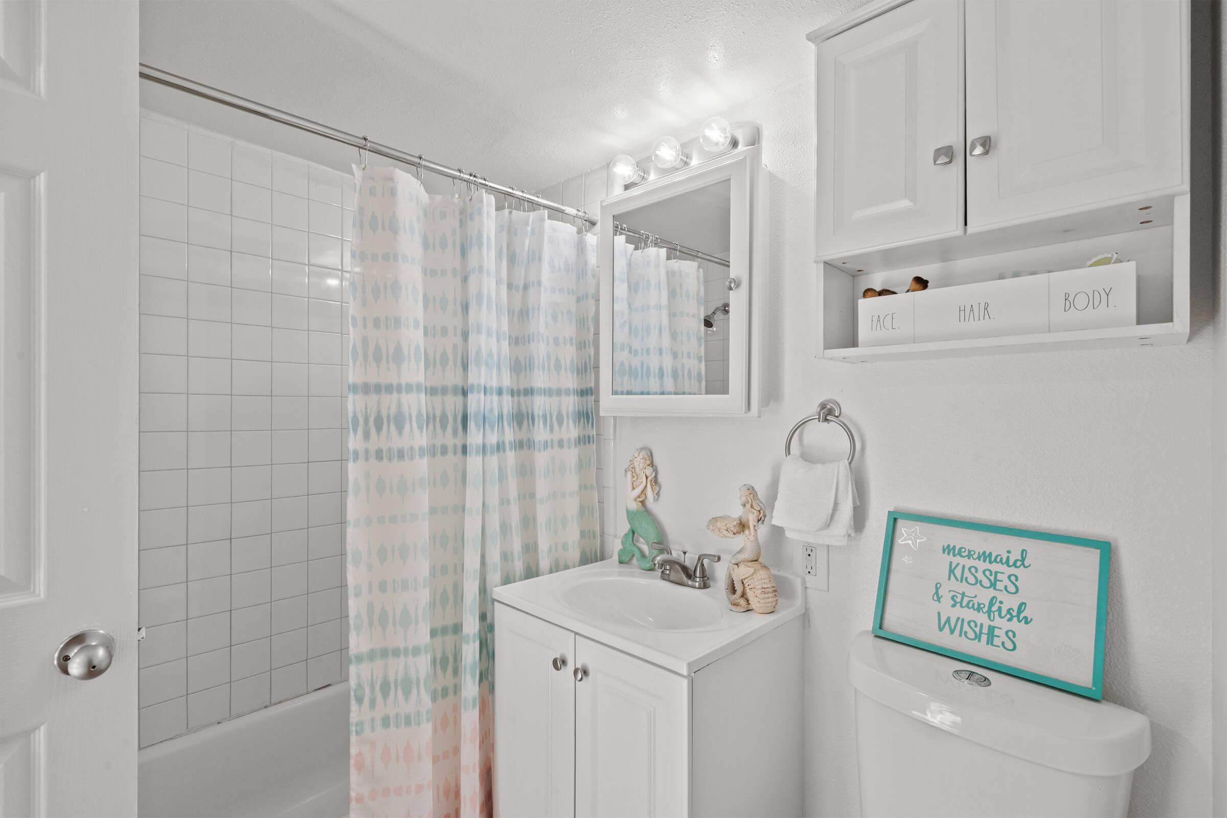 A bright bathroom featuring a shower with a patterned curtain, a white vanity with a sink, and decorative items including a mermaid figurine. The wall has a small shelf with a sign that reads "mermaid KISSES & starfish WISHES." The overall decor is light and welcoming.