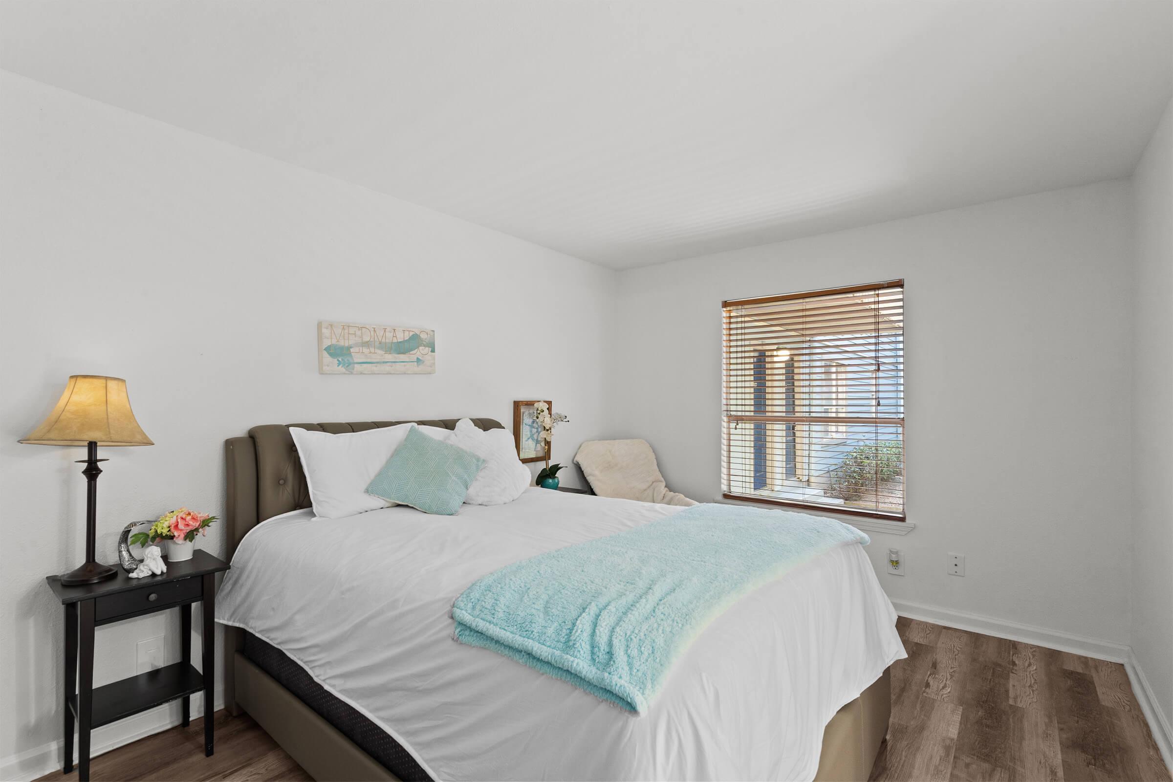 A cozy bedroom featuring a large bed with a white duvet and a light blue throw. There's a bedside table with a lamp and a small vase of flowers. A comfy chair is positioned next to a window with wooden blinds, allowing natural light to fill the room. The walls are painted white, creating a bright and airy atmosphere.