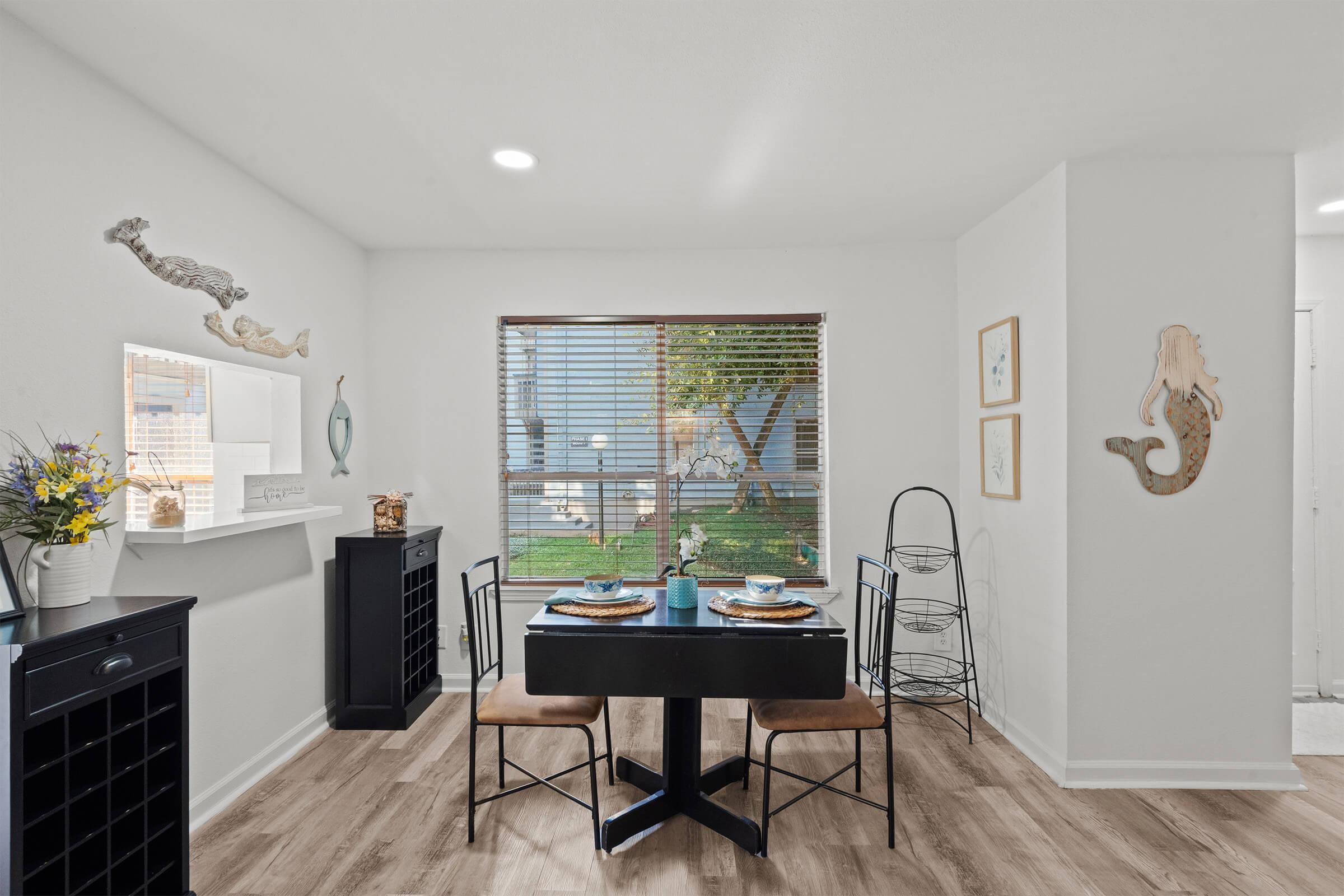 A cozy dining area featuring a black table and chairs set for two, surrounded by white walls adorned with decorative pieces. Natural light streams through a window, highlighting a small shelf with flowers and a patterned rug on the floor. A charming atmosphere with a touch of seaside decor.