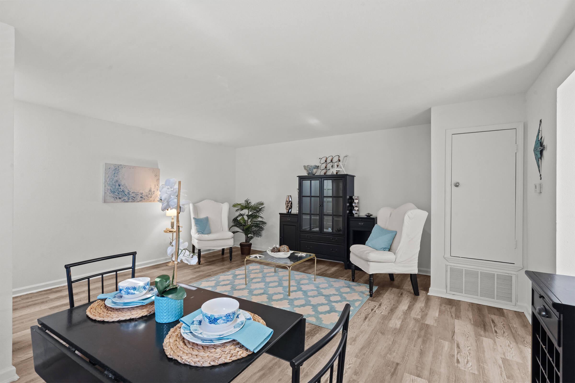 A bright, modern living room featuring a black dining table set with blue plates, woven placemats, and a decorative centerpiece. The space includes two white armchairs, a black cabinet, and a blue-patterned area rug on light wood flooring, complemented by minimal decor and a potted plant.