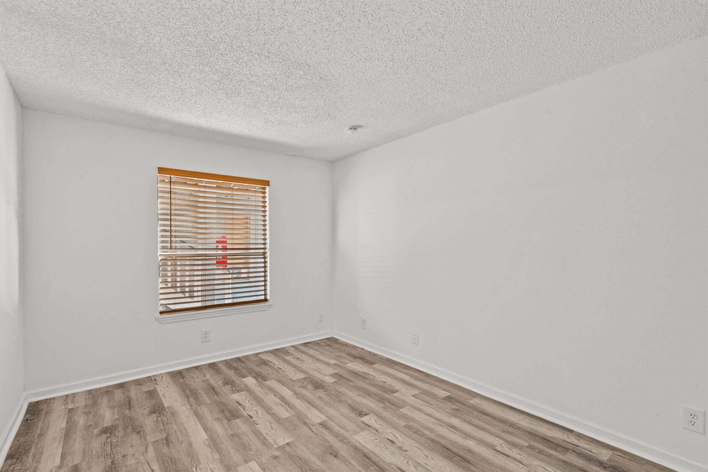 Empty room with light-colored walls and hardwood flooring. A window with blinds allows natural light inside, and there are no furnishings in the space. The atmosphere is bright and minimalistic, suitable for various uses like an office or bedroom.