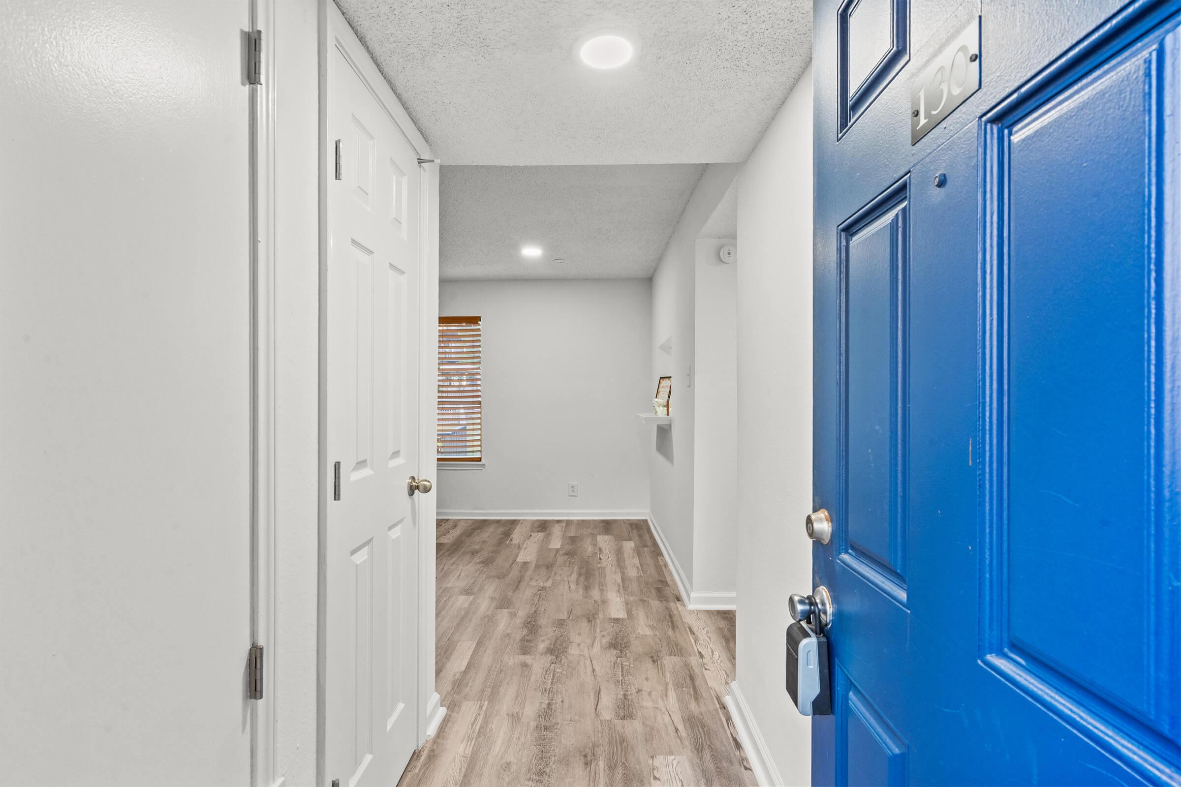 A bright entryway featuring a blue front door, two white closet doors, and light-colored wood flooring. The walls are painted white, and there is a small shelf along one side. Natural light enters from a window with blinds in the background.
