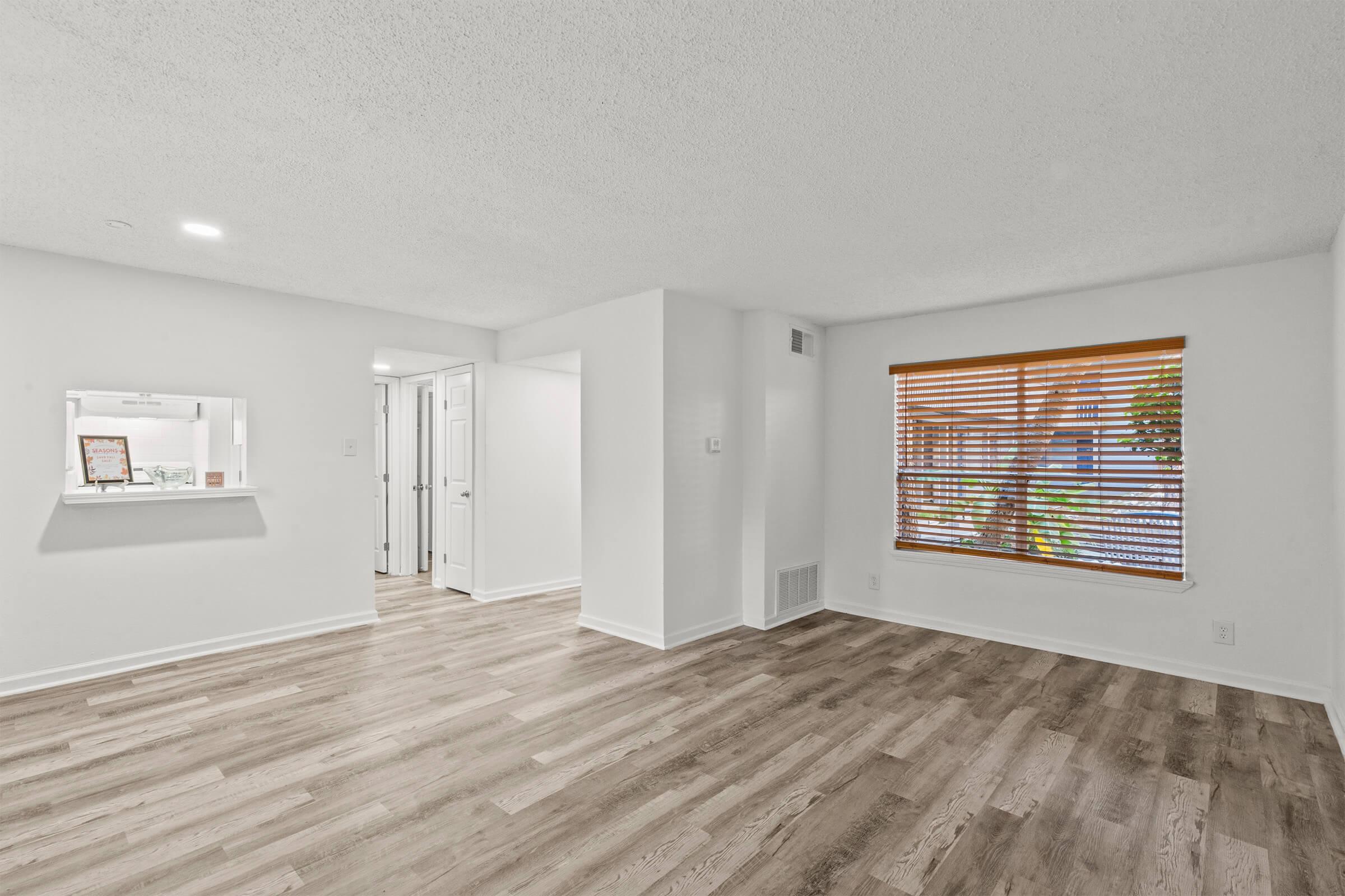 Spacious room with light-colored walls and a textured ceiling. The wooden floor adds warmth, and a large window with wooden blinds allows natural light to filter in. A small opening in the wall leads to another room, enhancing the open layout of the space.
