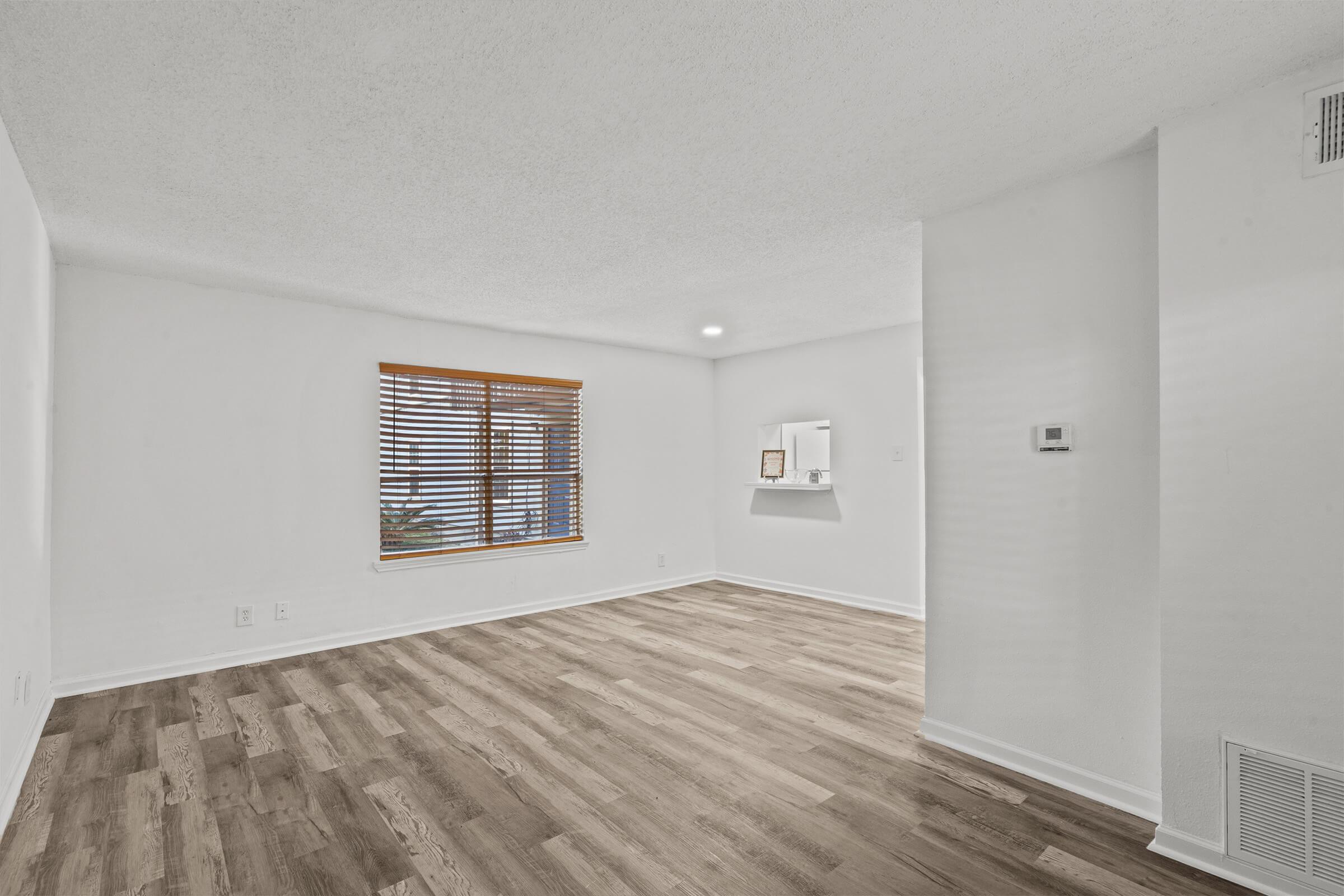 Spacious interior of a room with light-colored walls and wooden flooring. Natural light pours in through a window with wooden blinds. There is a small wall-mounted shelf and a thermostat on the adjacent wall, creating a bright, airy atmosphere.