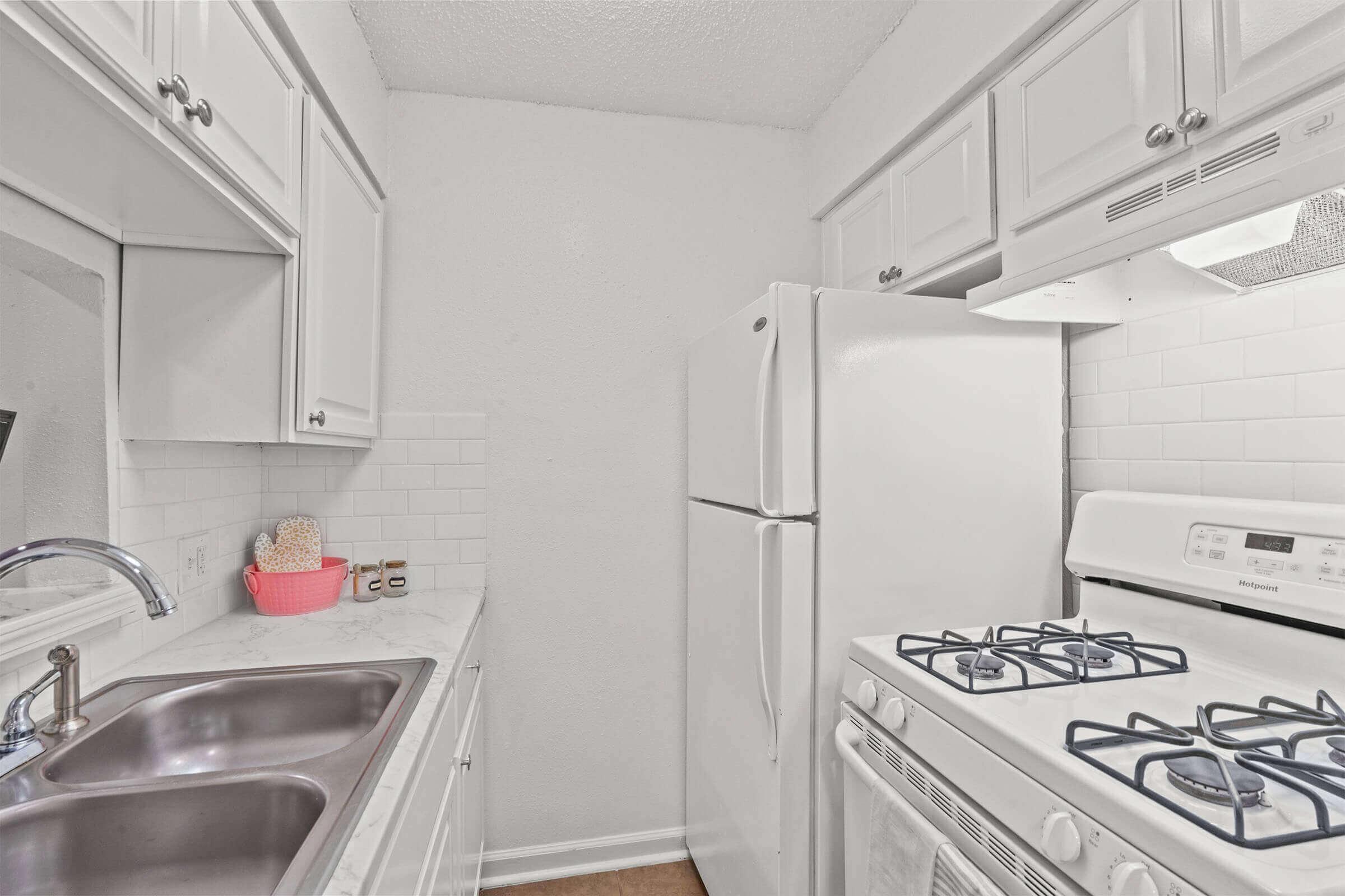 A small, clean kitchen featuring white cabinets, a double sink, and stainless steel appliances. The refrigerator is white, and a gas stove is visible. Light-colored countertops and a tiled backsplash add to the bright, modern appearance. Bright lighting enhances the spacious feel of the area.