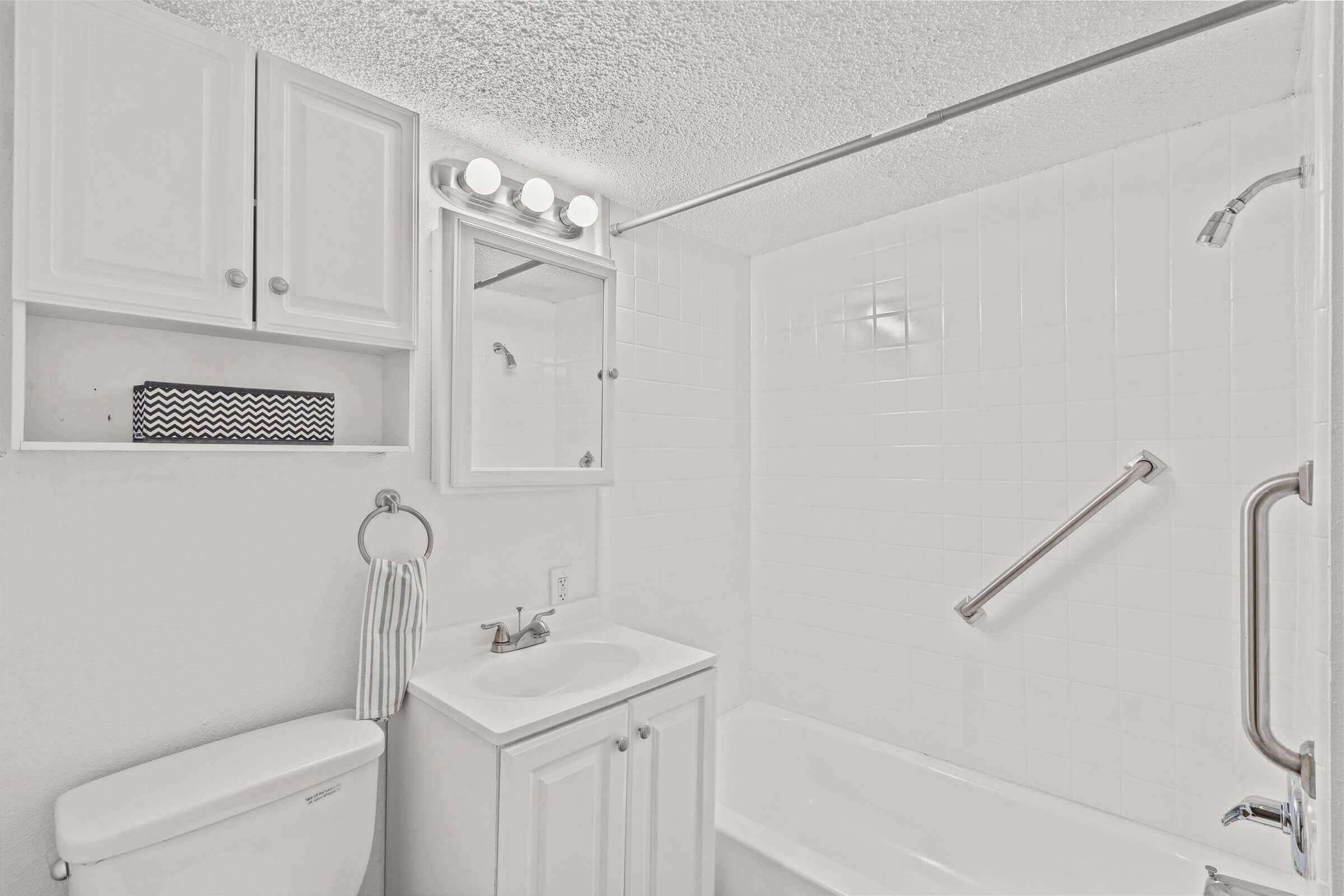 A bright and clean bathroom featuring a white bathtub, sink with cabinets, and mirror. A towel holder and a grab bar are visible, along with a light fixture above the mirror. The walls are tiled in white, contributing to a fresh, spacious atmosphere.